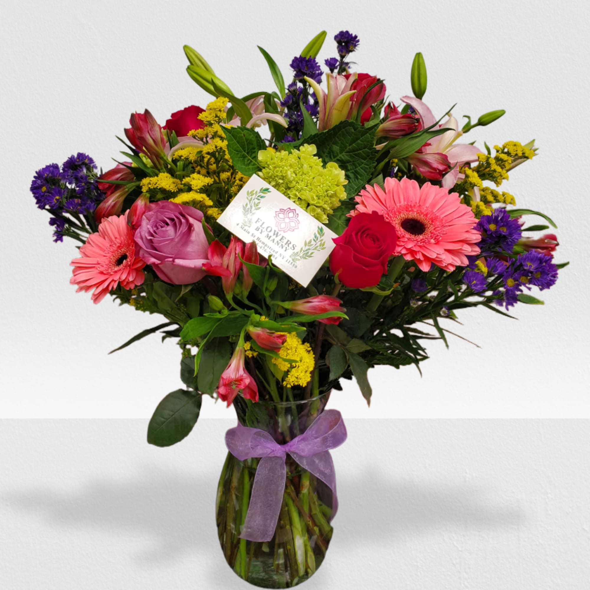 Mixed bouquet of pink gerberas, roses, lilies and purple accents in a glass vase with ribbon