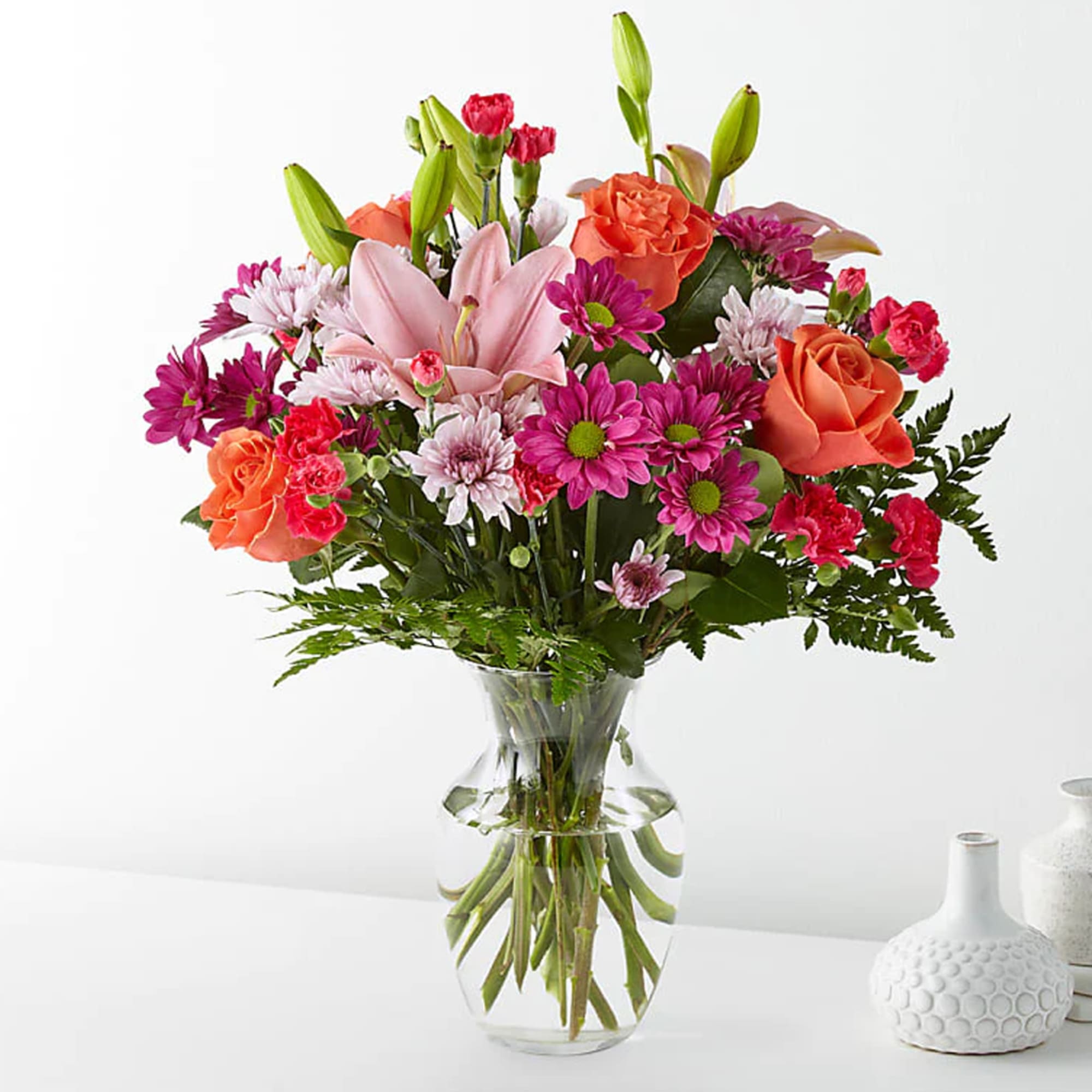 Mixed bouquet of pink lilies, orange roses, and magenta daisies in a clear glass vase