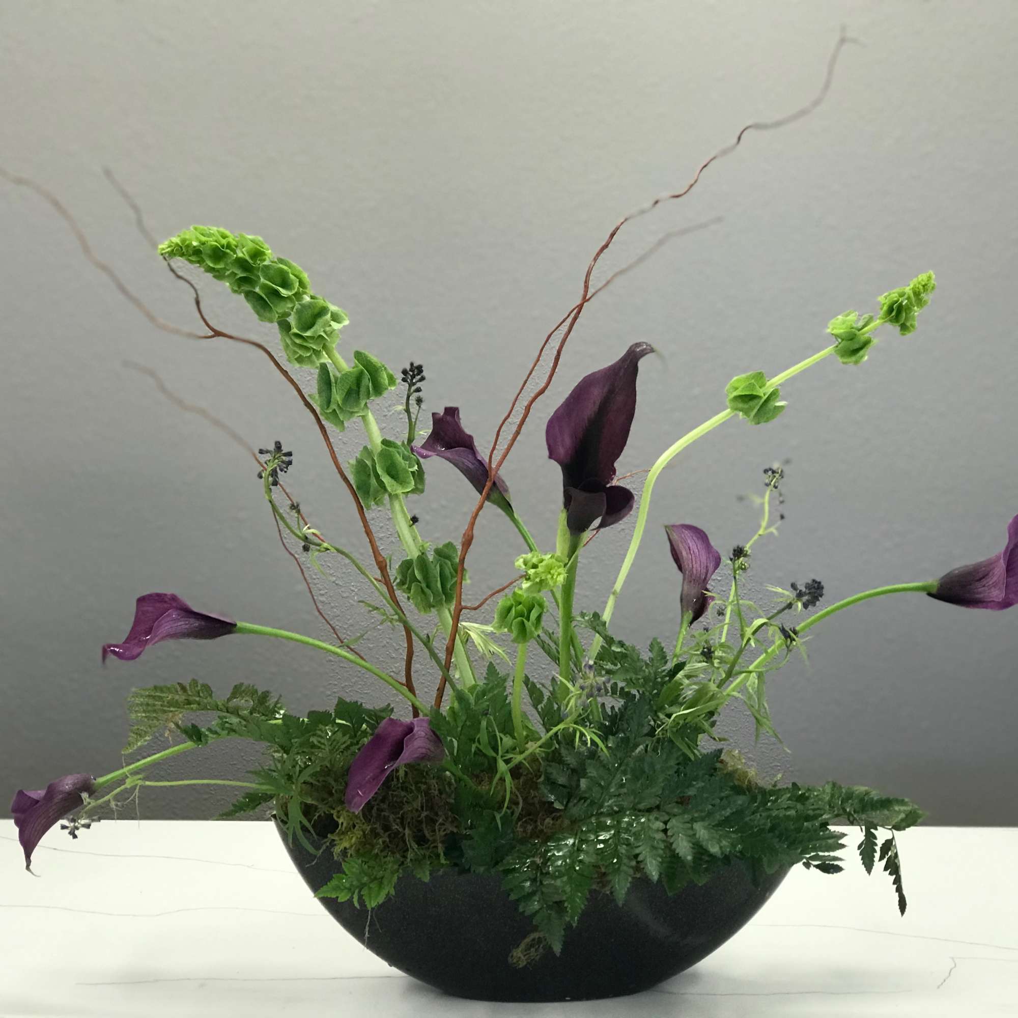 Purple calla lilies in a low black bowl with green foliage