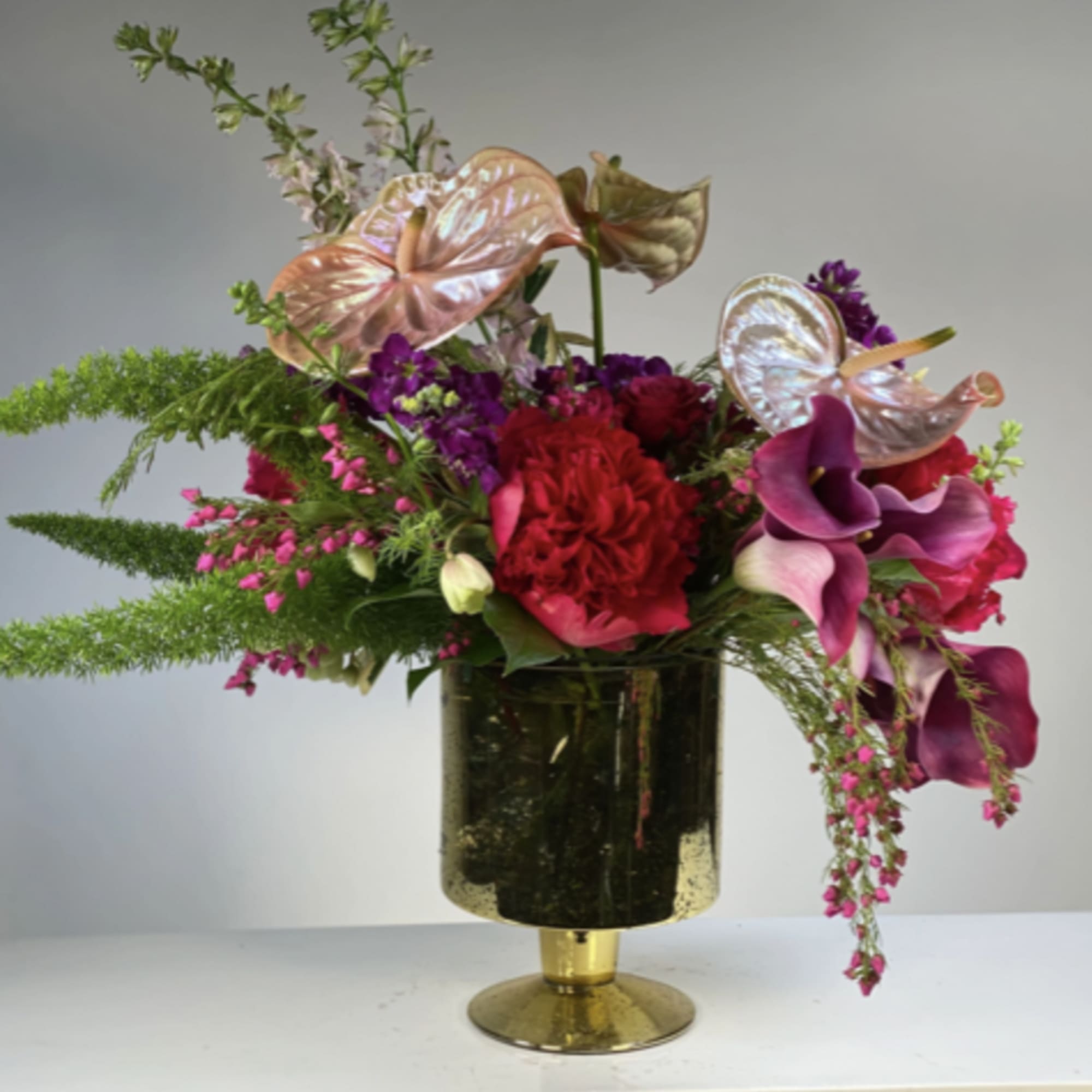Asymmetrical arrangement of red and magenta flowers with calla lilies in a gold pedestal vase.