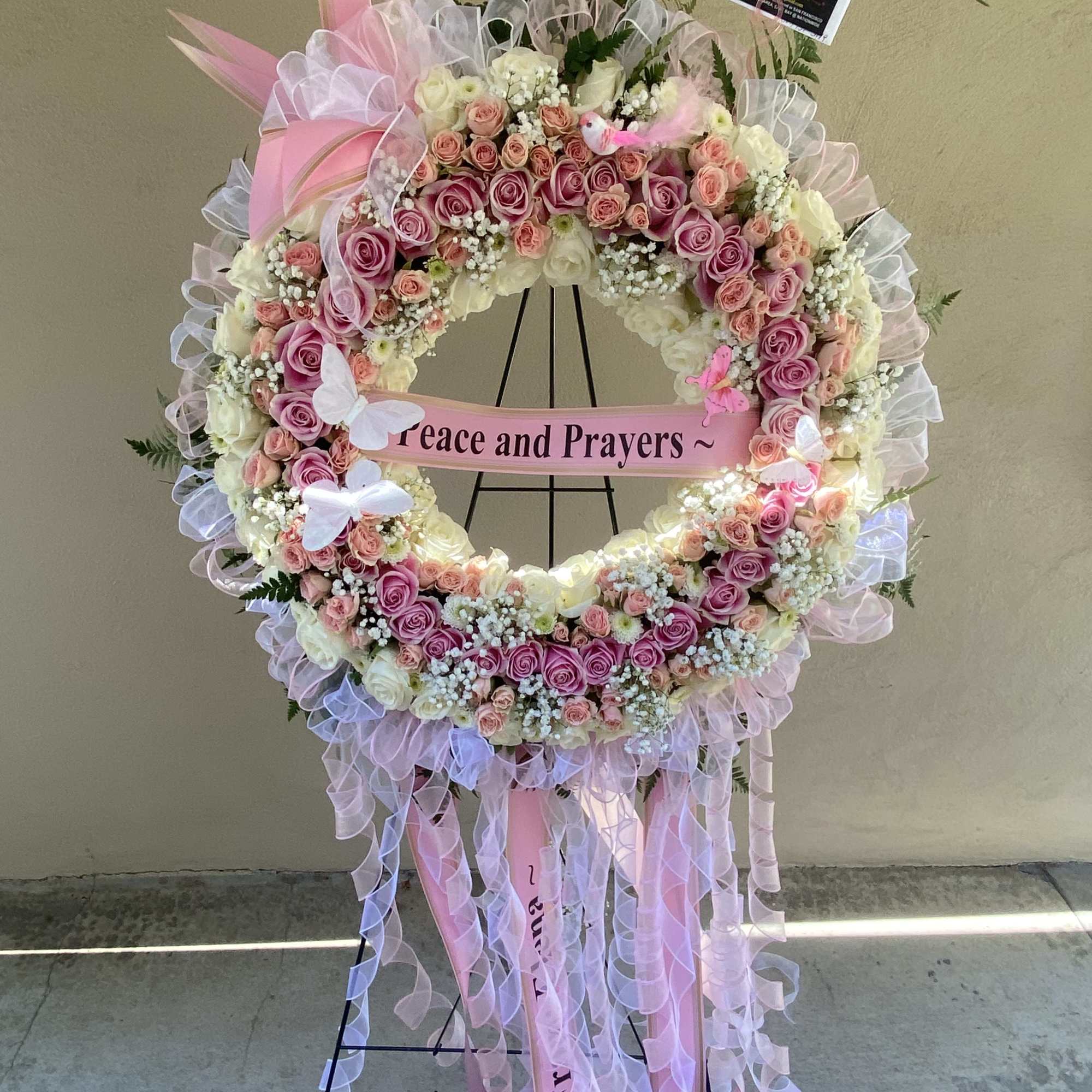 Pink and white rose wreath on a stand with a "Peace and Prayers" ribbon