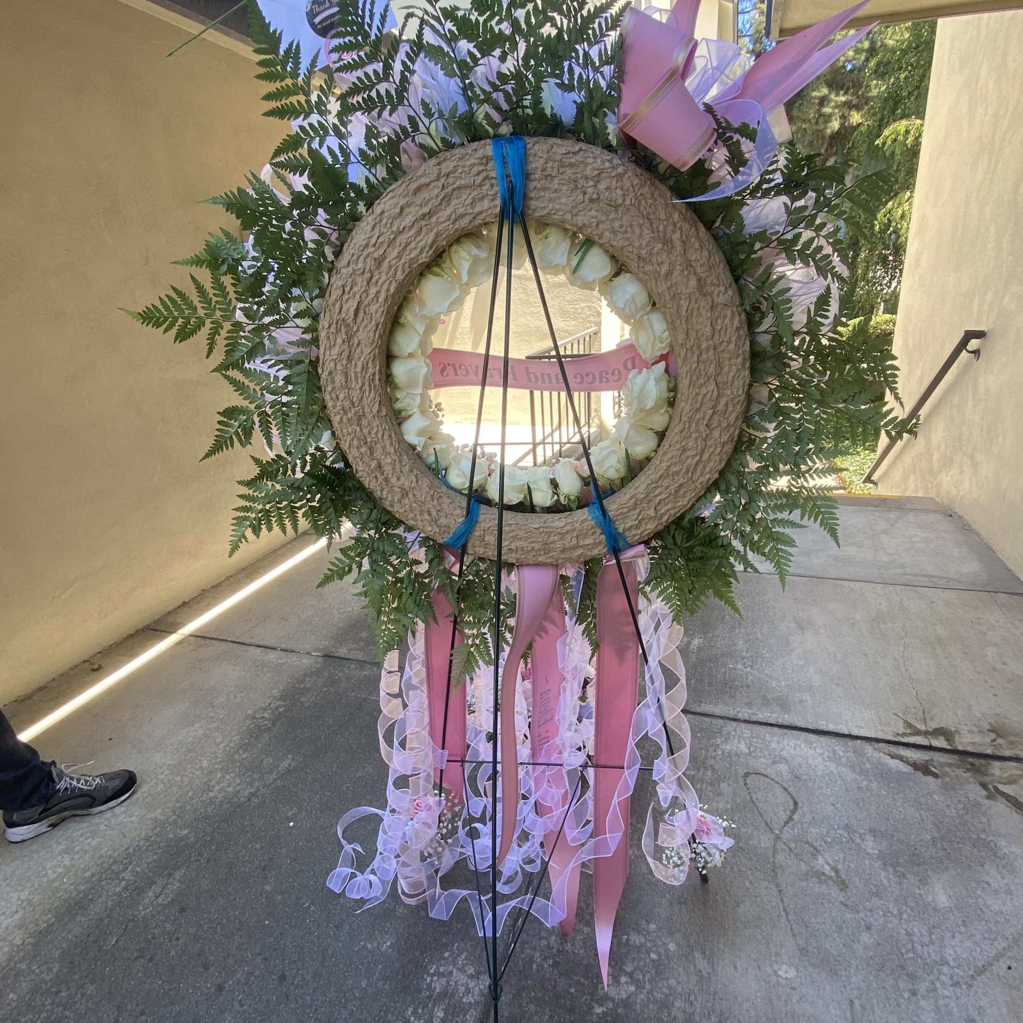 Large floral wreath with white roses, pink ribbons, and fern greenery on a stand