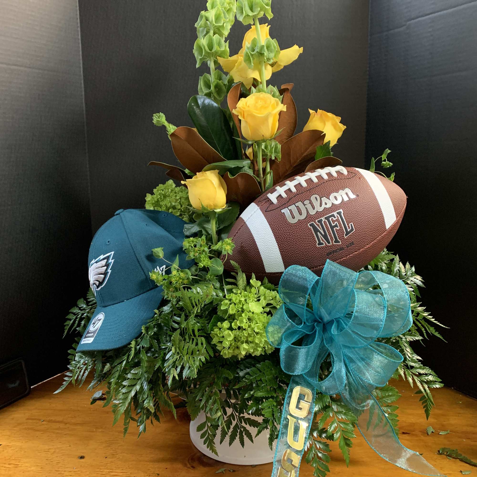 Yellow roses arranged with a football, cap, and teal ribbon in a white container
