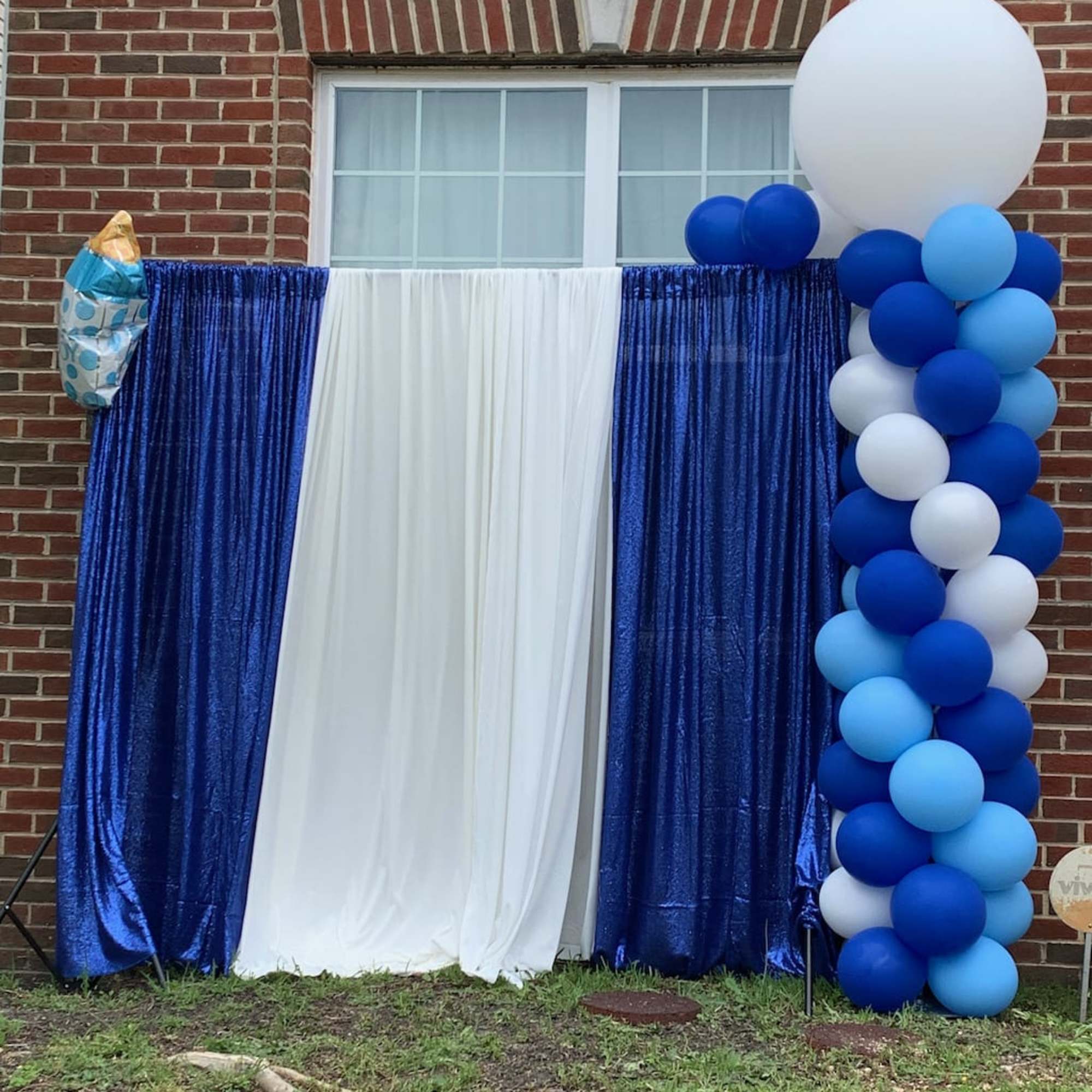 Blue and white balloon arch with draped fabric backdrop against a brick wall