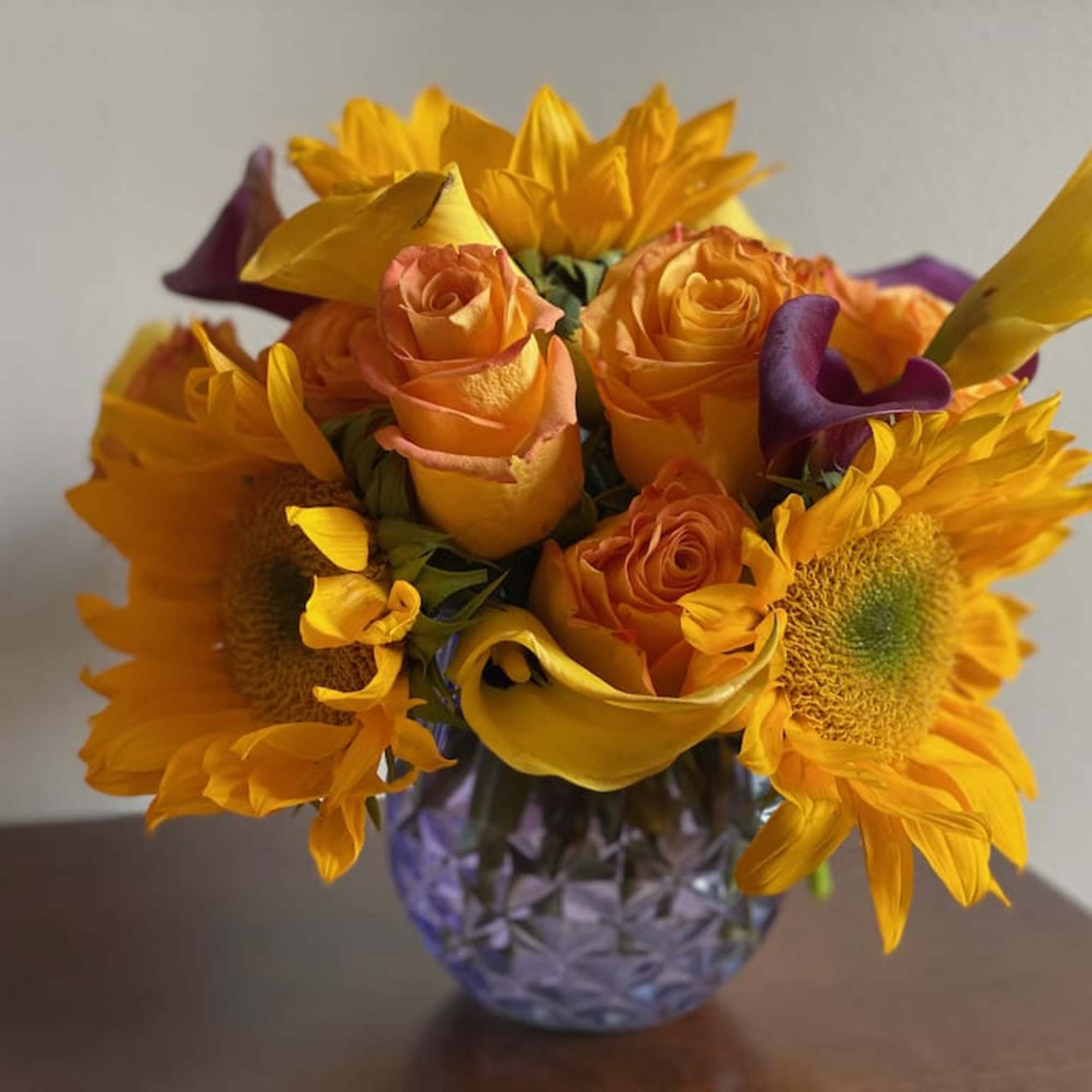 Bouquet of orange roses, sunflowers, and purple calla lilies in a purple vase