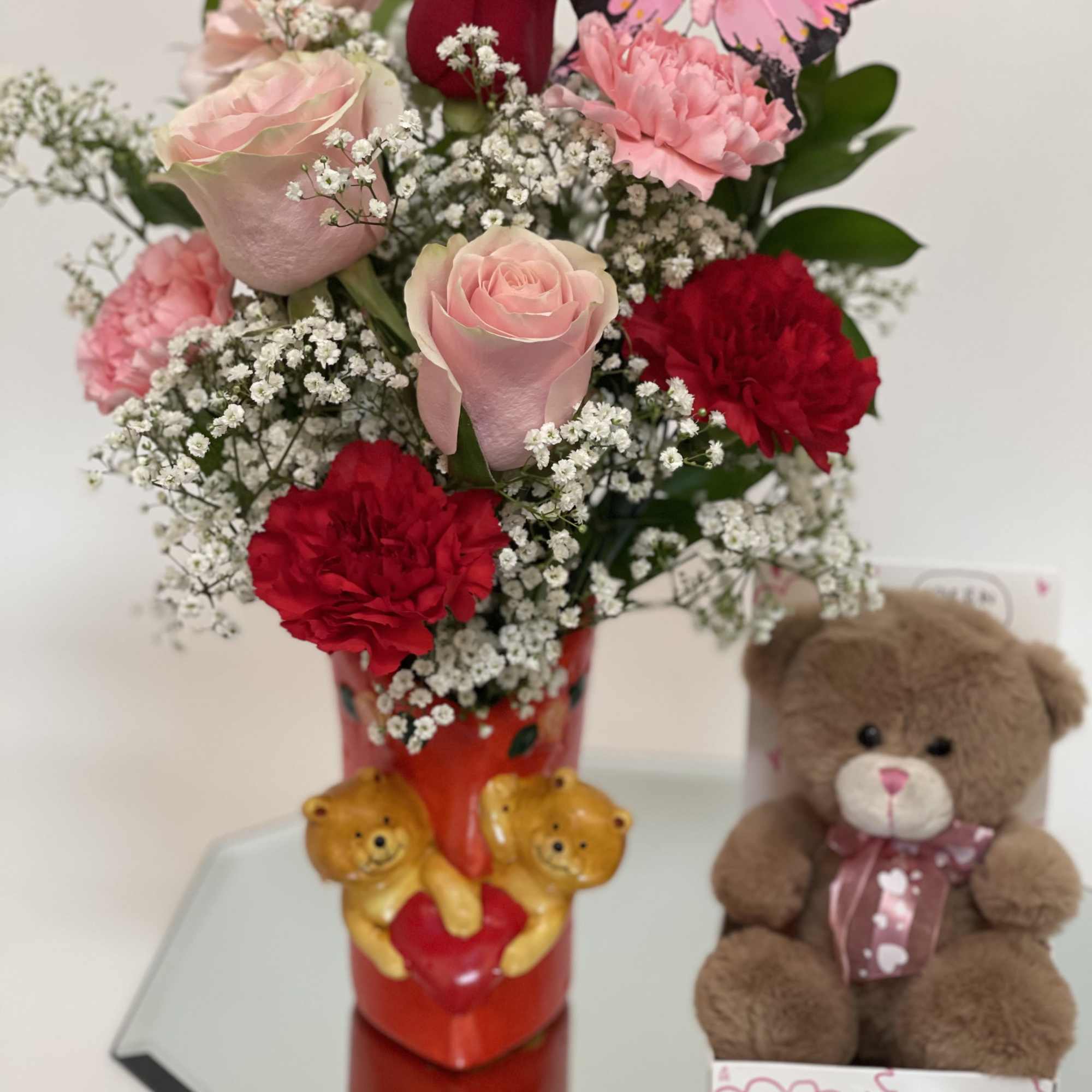 Bouquet of pink and red roses with carnations in a red vase, beside a teddy bear
