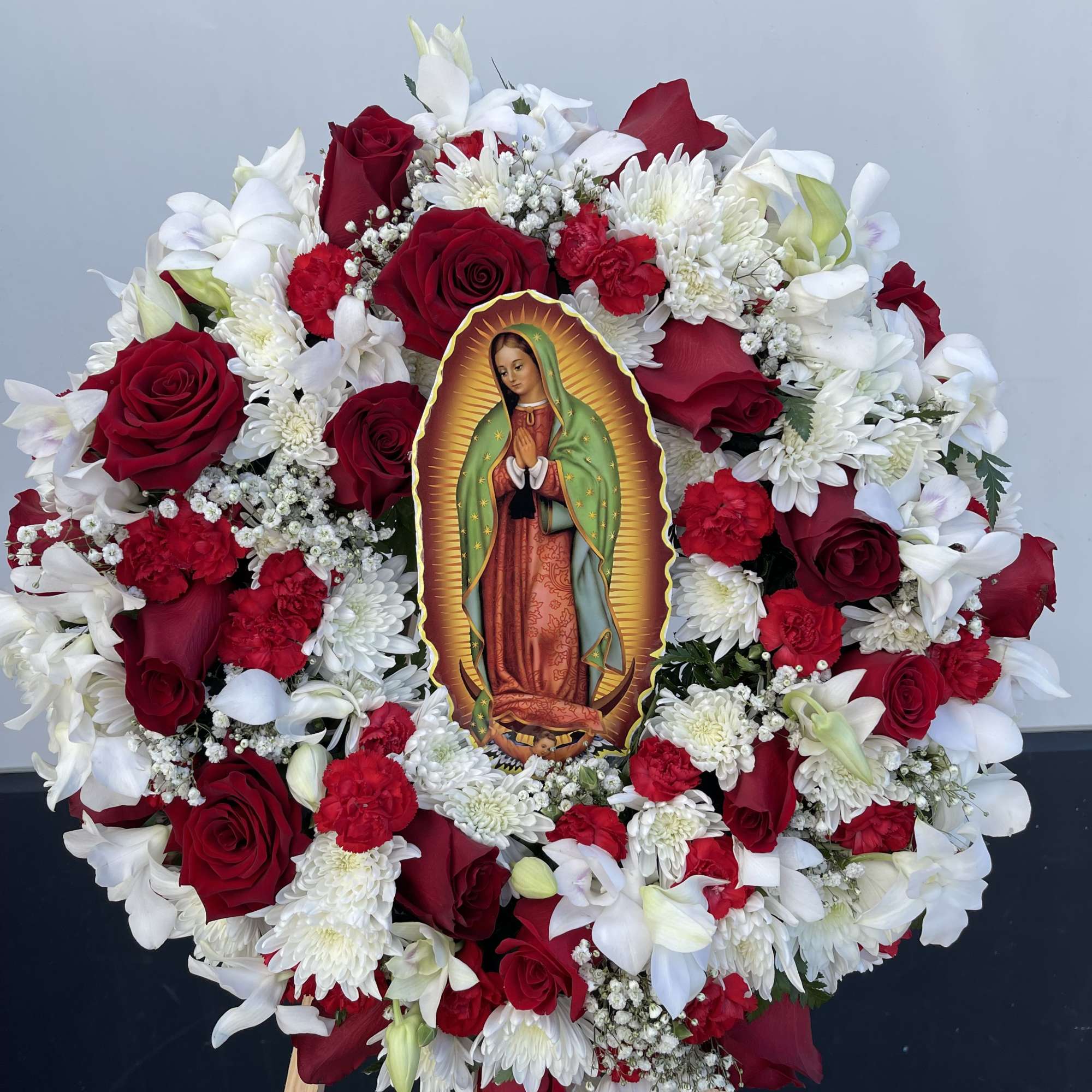 Circular floral wreath with red roses, white flowers, and a Virgin of Guadalupe image