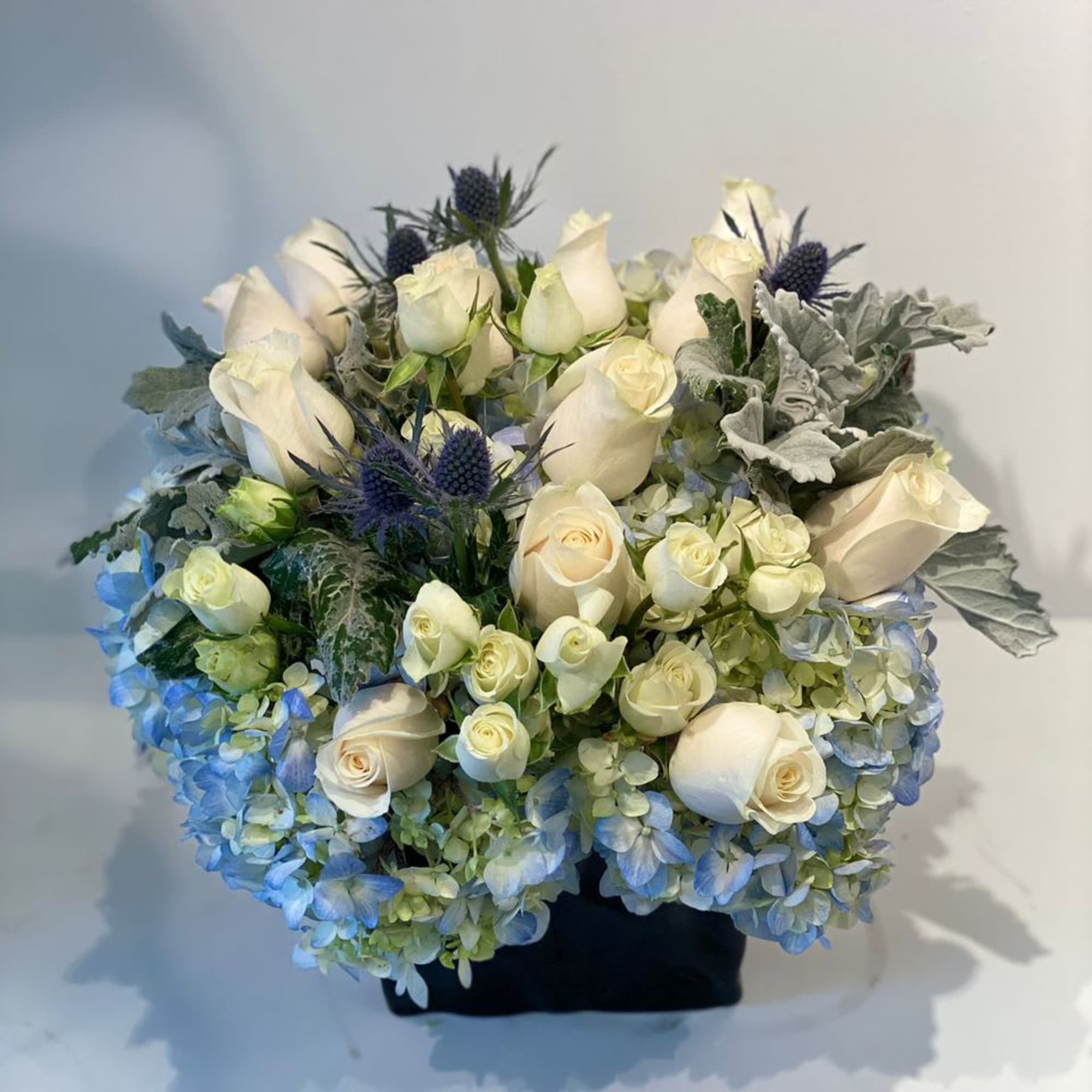 Bouquet of white roses, blue hydrangeas, and thistle in a black container