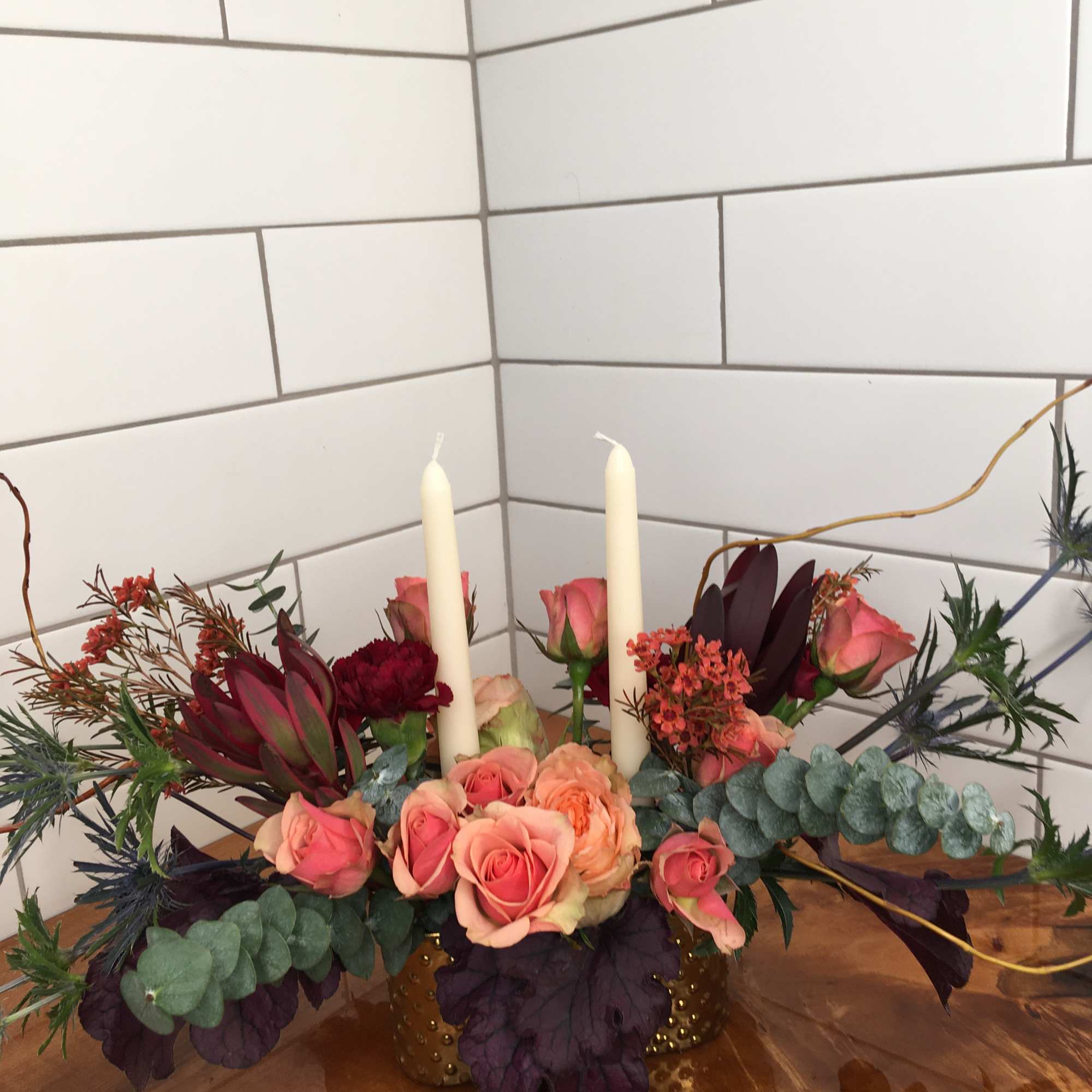 Pink roses and burgundy flowers arranged with two white candles in a gold vase