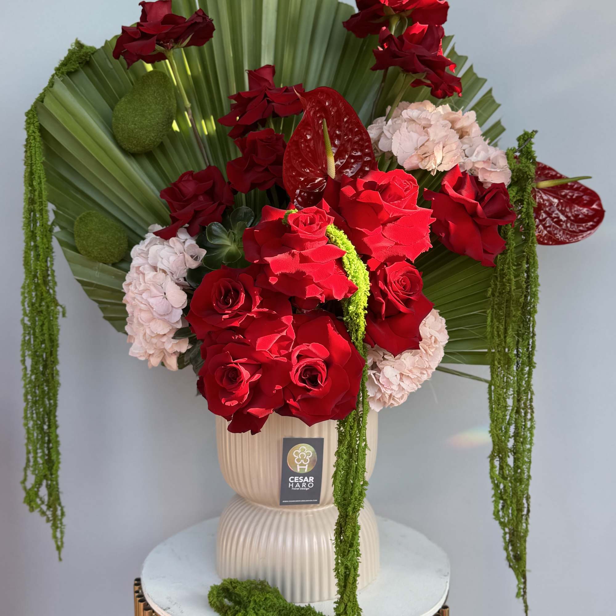 A dramatic white vase arrangement with palms, red roses, anthuriums, pink hydrangeas