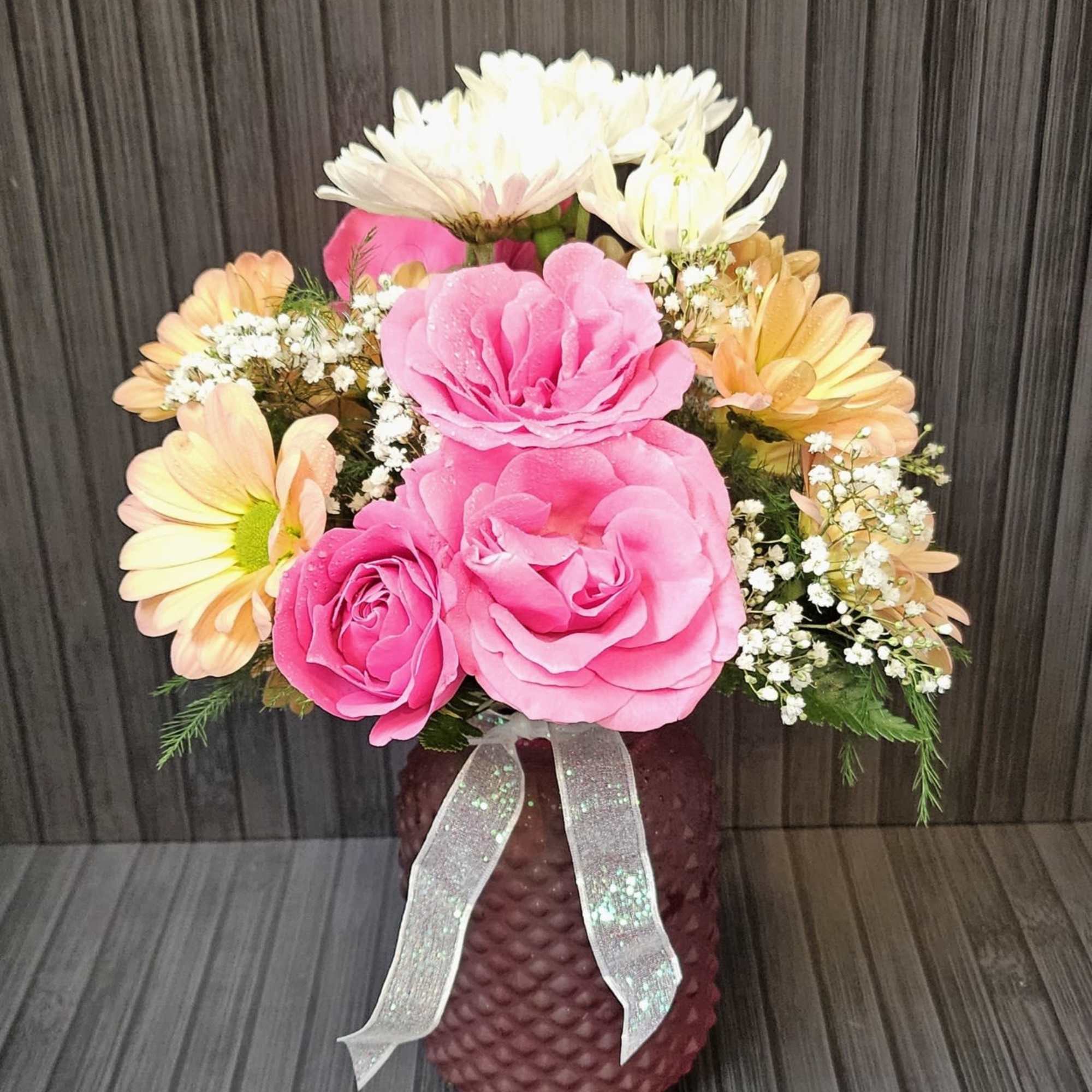 Pink roses and peach daisies arranged in a textured vase with a ribbon.