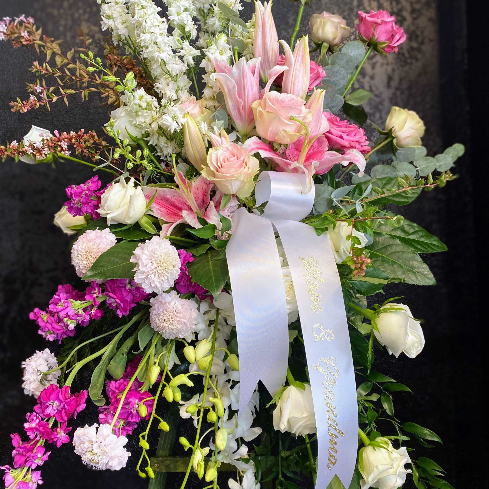 Tall standing spray of pink lilies, roses, and white flowers with a white ribbon on an easel.