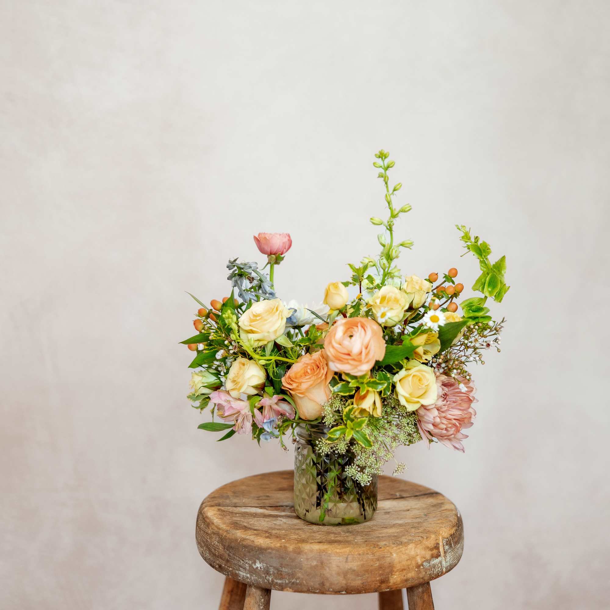 Mixed pastel flower arrangement in a textured glass vase on a wooden stool