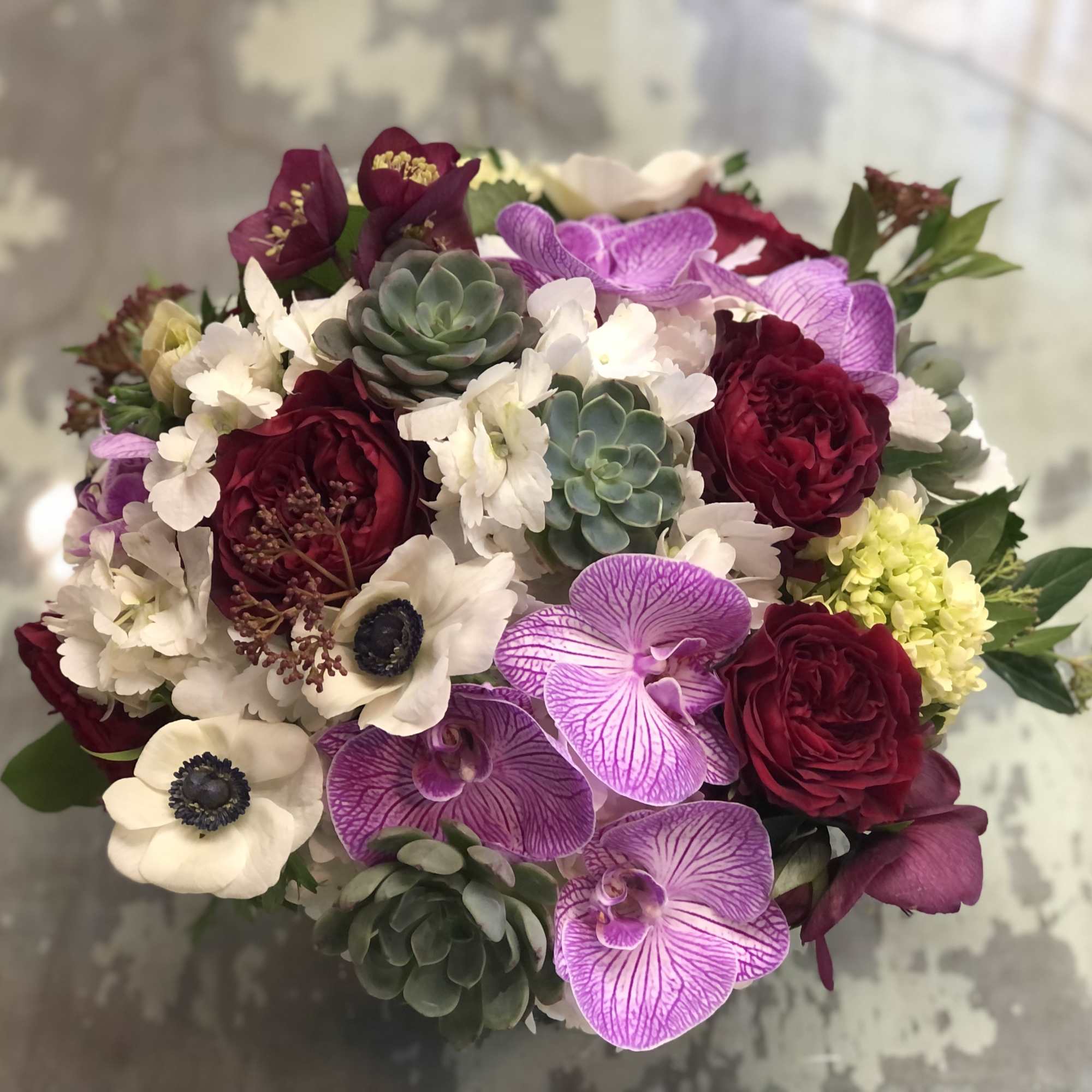 Round bouquet with burgundy roses, purple orchids, white anemones, and succulents