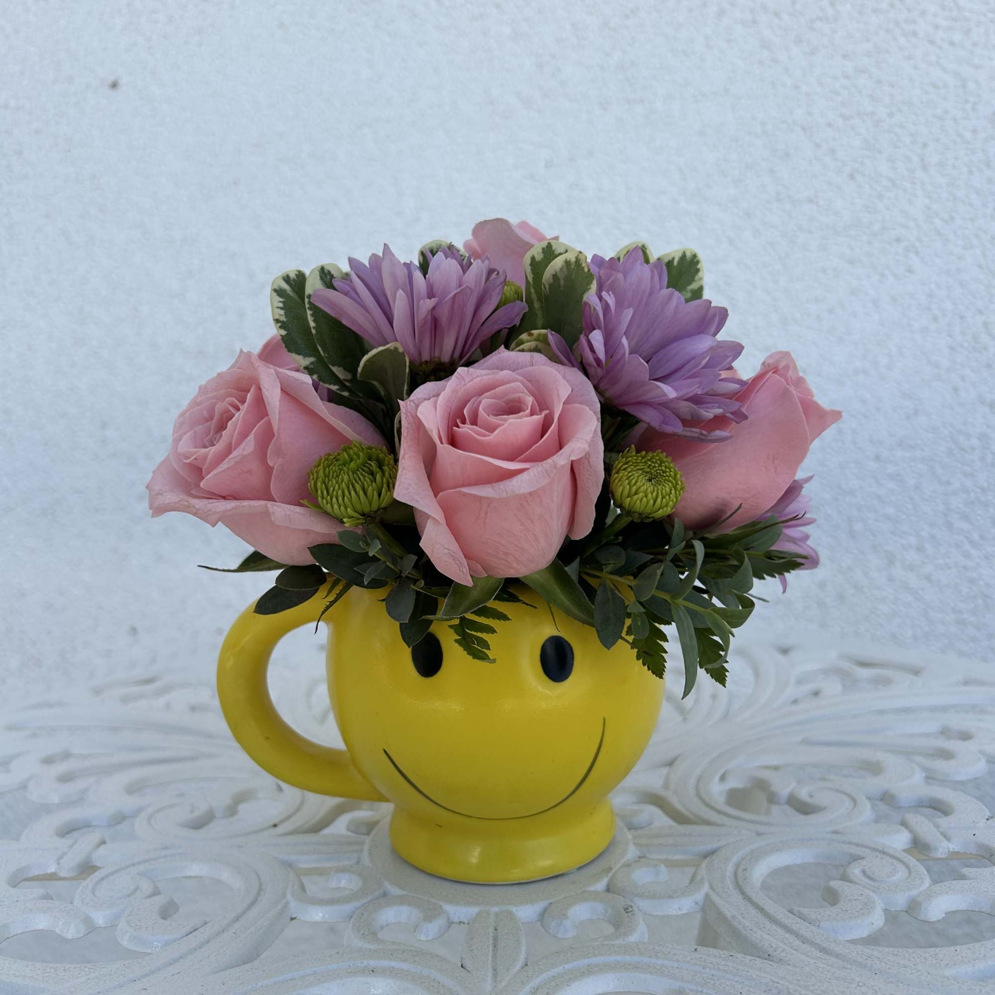Pink roses and purple daisies arranged in a yellow smiley-face mug