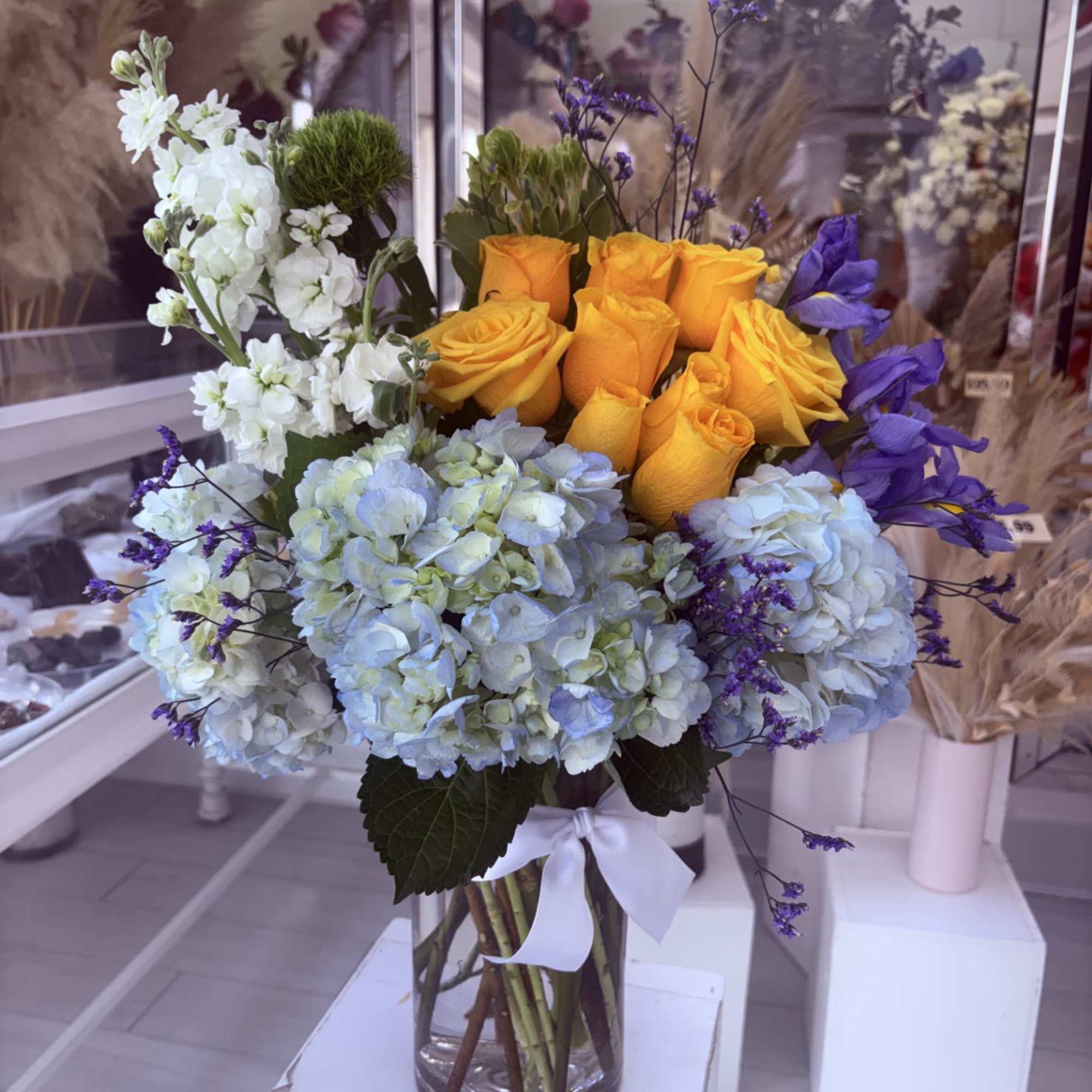 Vase arrangement of yellow roses, blue hydrangeas, white blooms and purple accents in a clear glass cylinder
