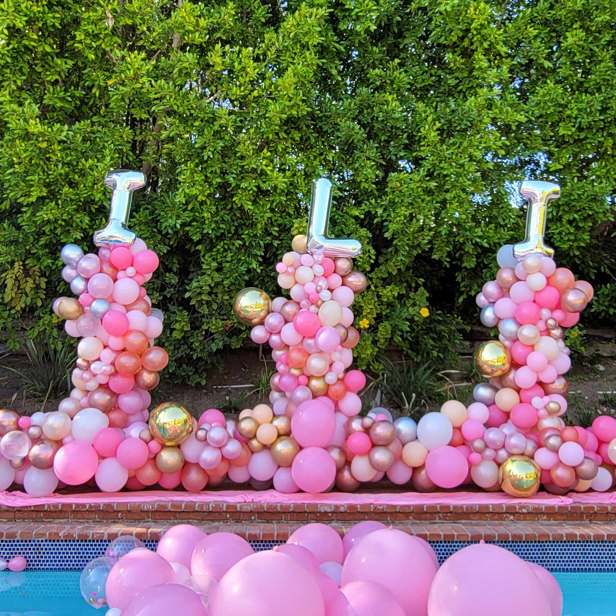 Pink balloon display spelling "LILIA" outdoors by a pool