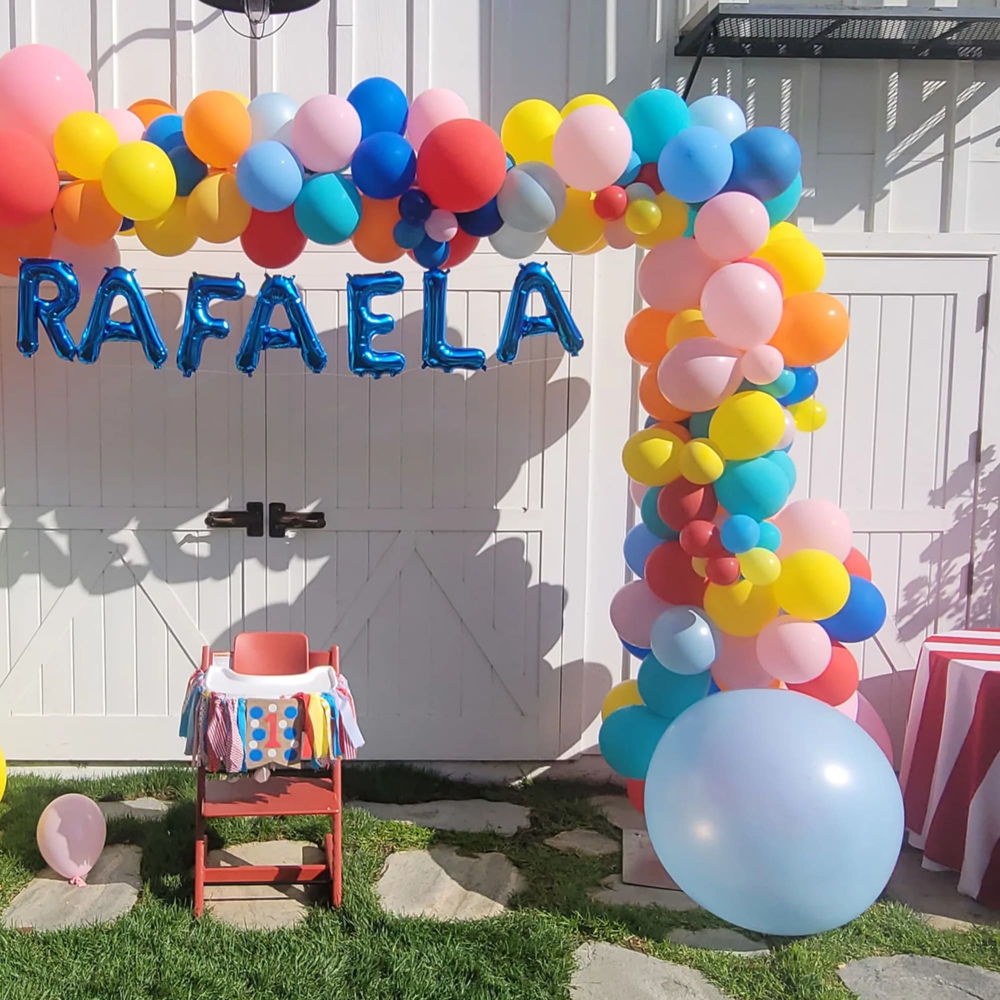 Colorful balloon arch with blue "RAFAELA" letters and a small decorated chair
