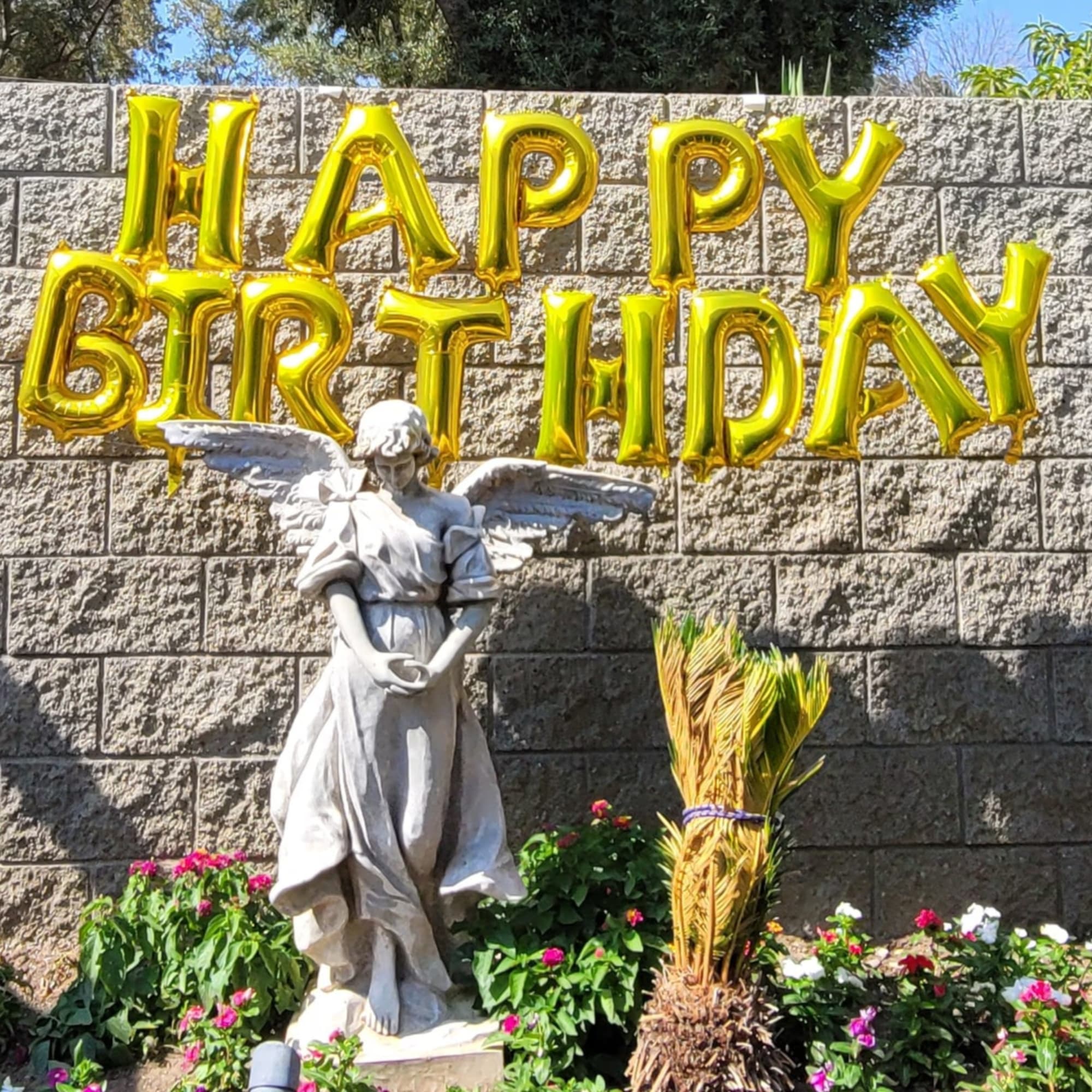 Gold balloon letters spelling Happy Birthday above a stone angel statue