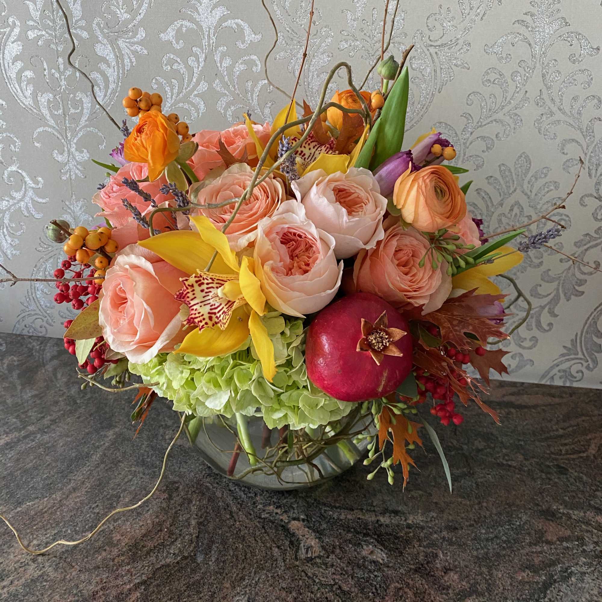 Mixed bouquet of peach roses, yellow orchids, and a pomegranate in a glass bowl