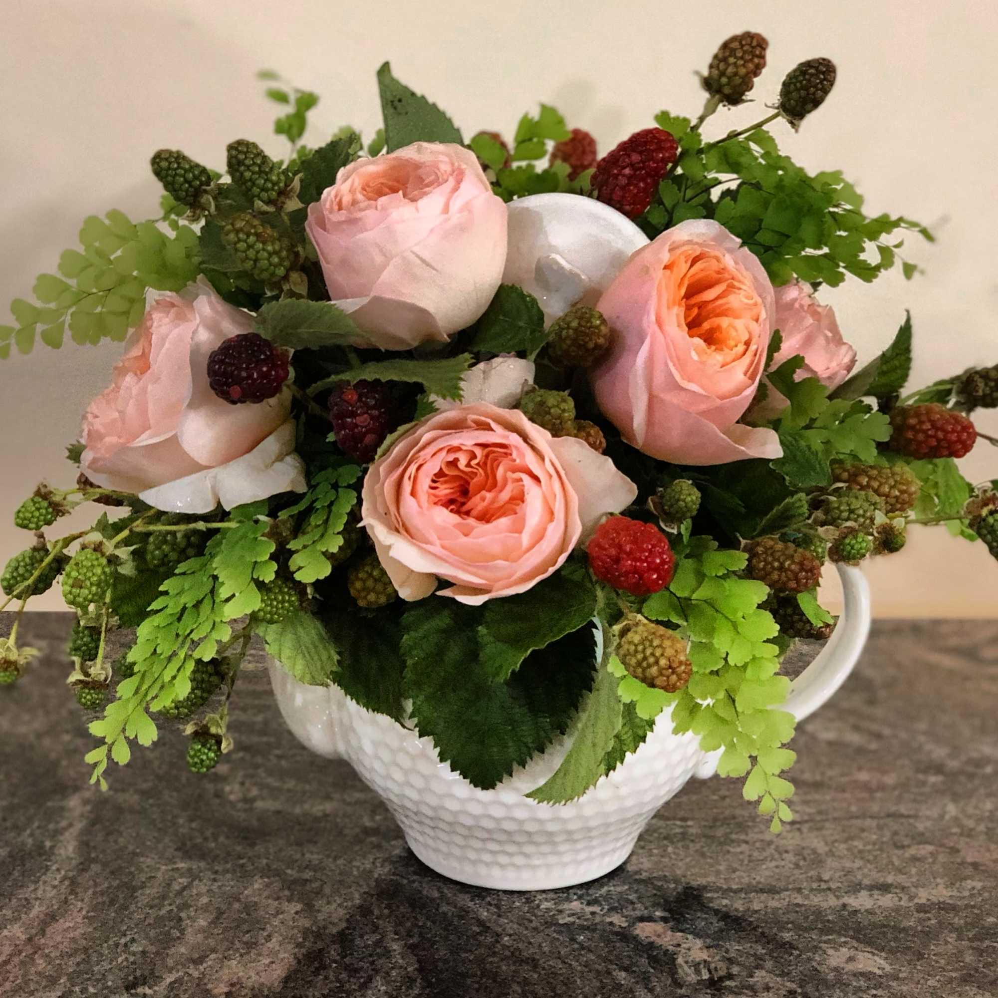 Pink roses arranged in a white ceramic pitcher with green berries and fern-like foliage.