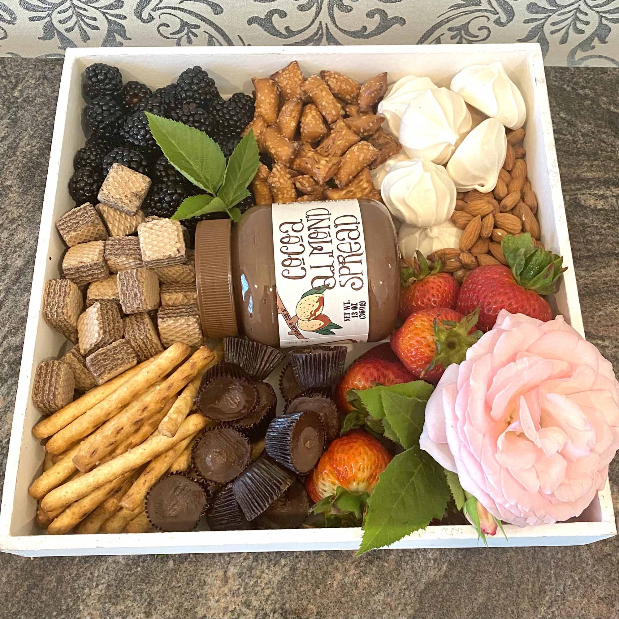 Gift tray with fruit, snacks, chocolate spread, and a pink artificial rose