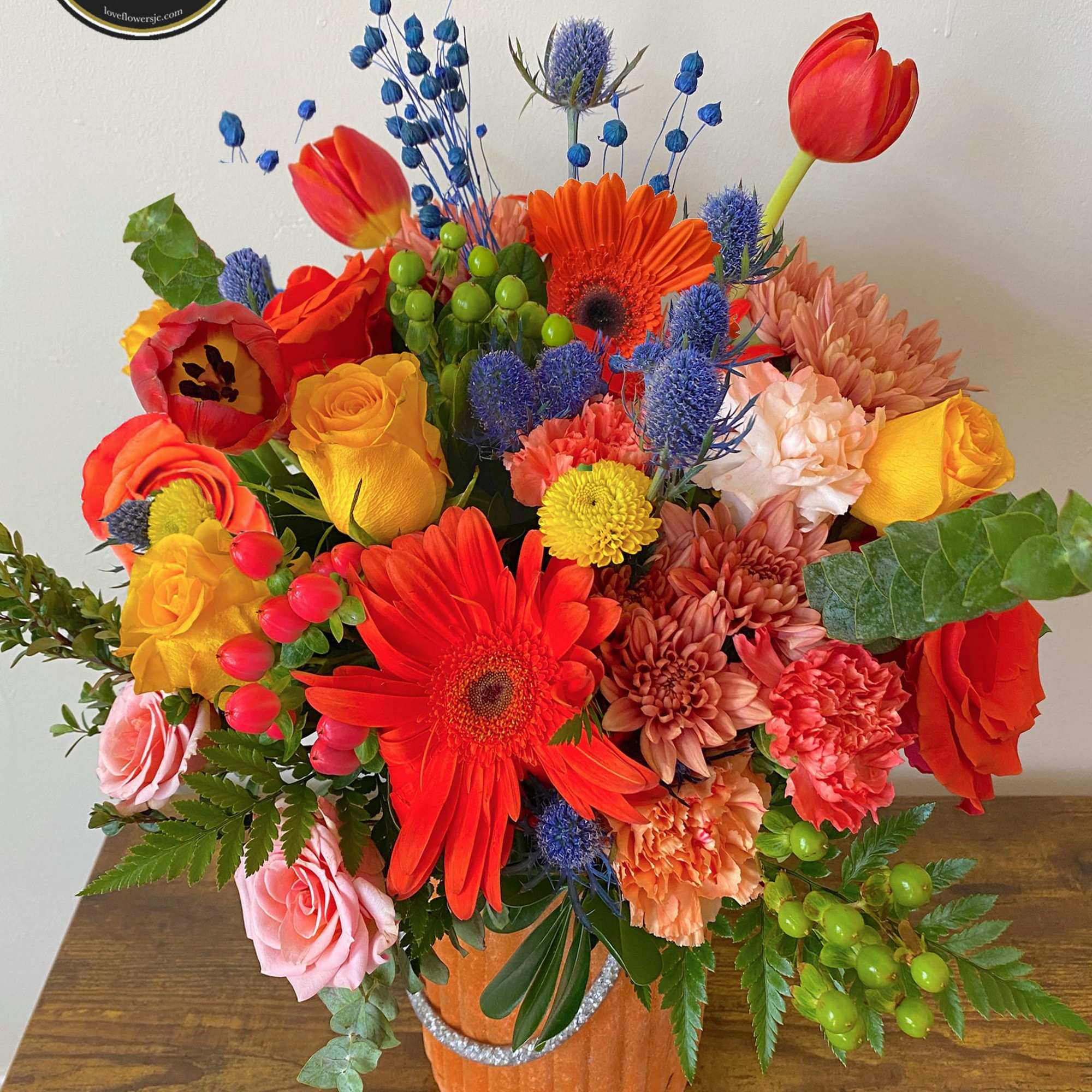 Bright mixed bouquet of orange gerbera daisies, roses, tulips and mums in an orange vase on a wooden table