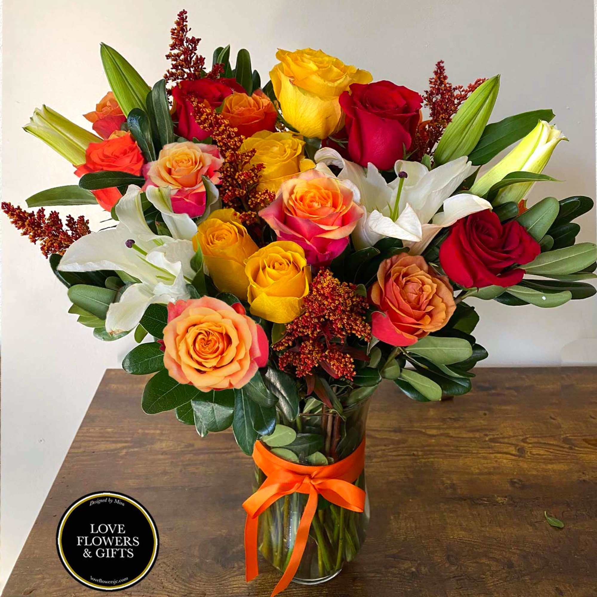 Tall vase arrangement of red, yellow, and orange roses with white lilies and an orange ribbon bow.