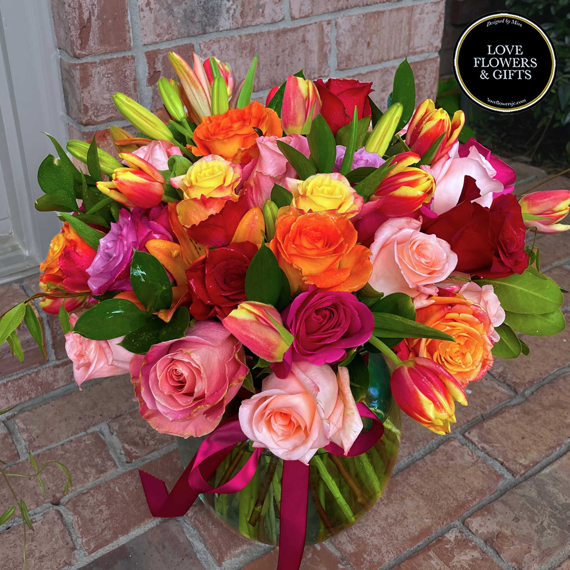 Bright bouquet of pink, orange, yellow, and red roses with tulips and lilies in a glass vase.