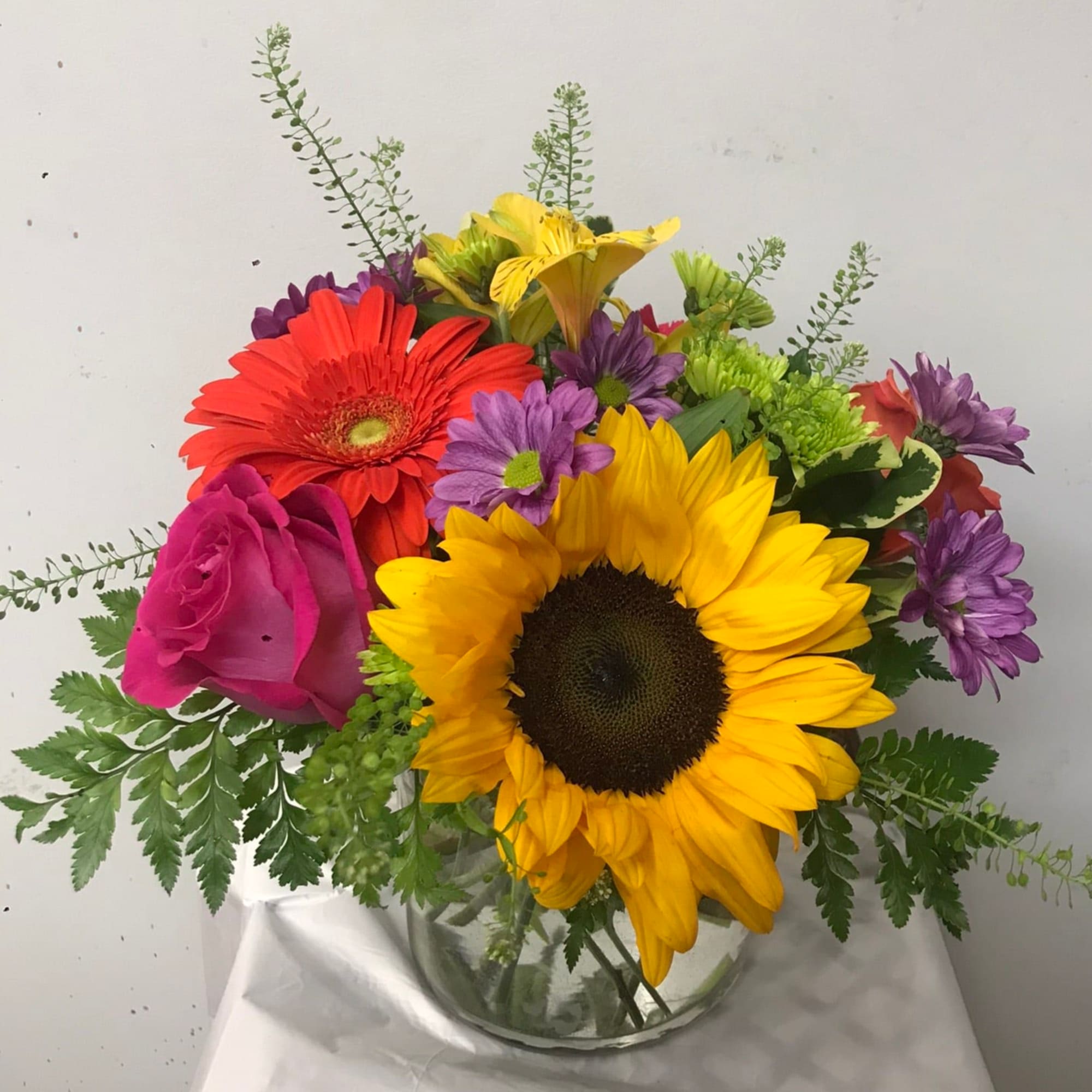 Mixed bouquet with sunflowers, roses, gerbera daisies, and purple blooms in a glass vase