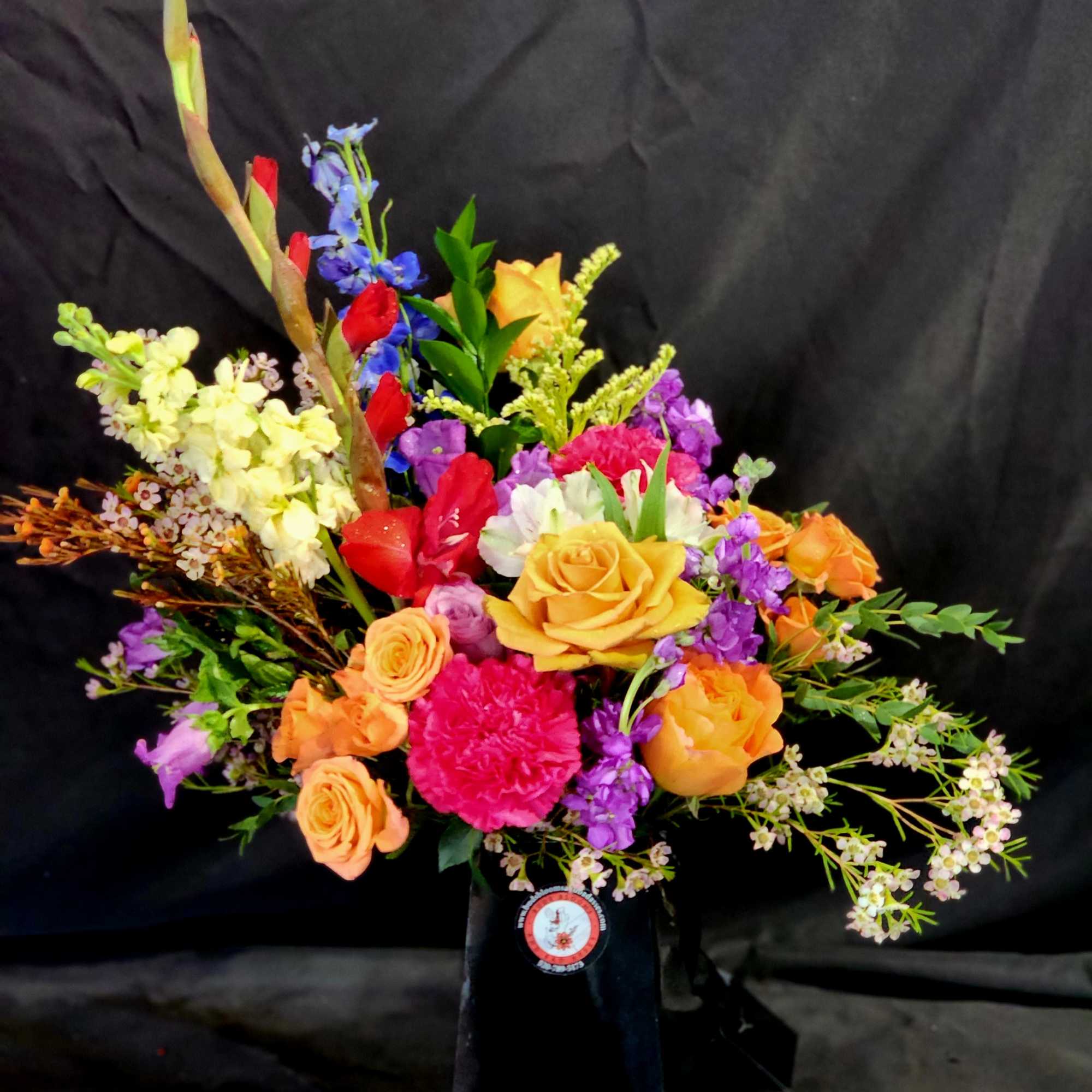 Tall mixed arrangement of orange roses, pink carnations, and assorted bright blooms in a black box vase