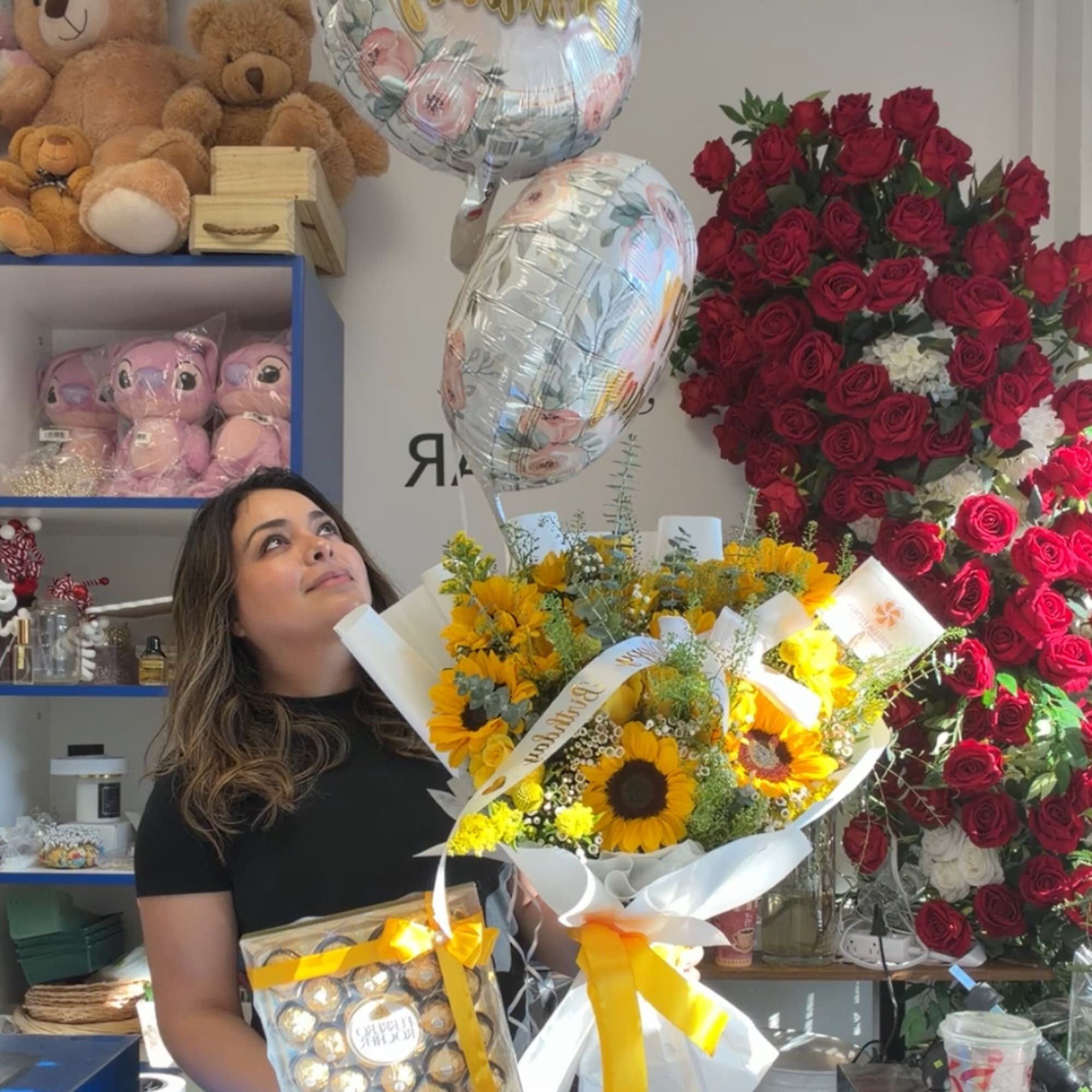 Woman holding a sunflower bouquet, chocolates, and birthday balloons in a flower shop