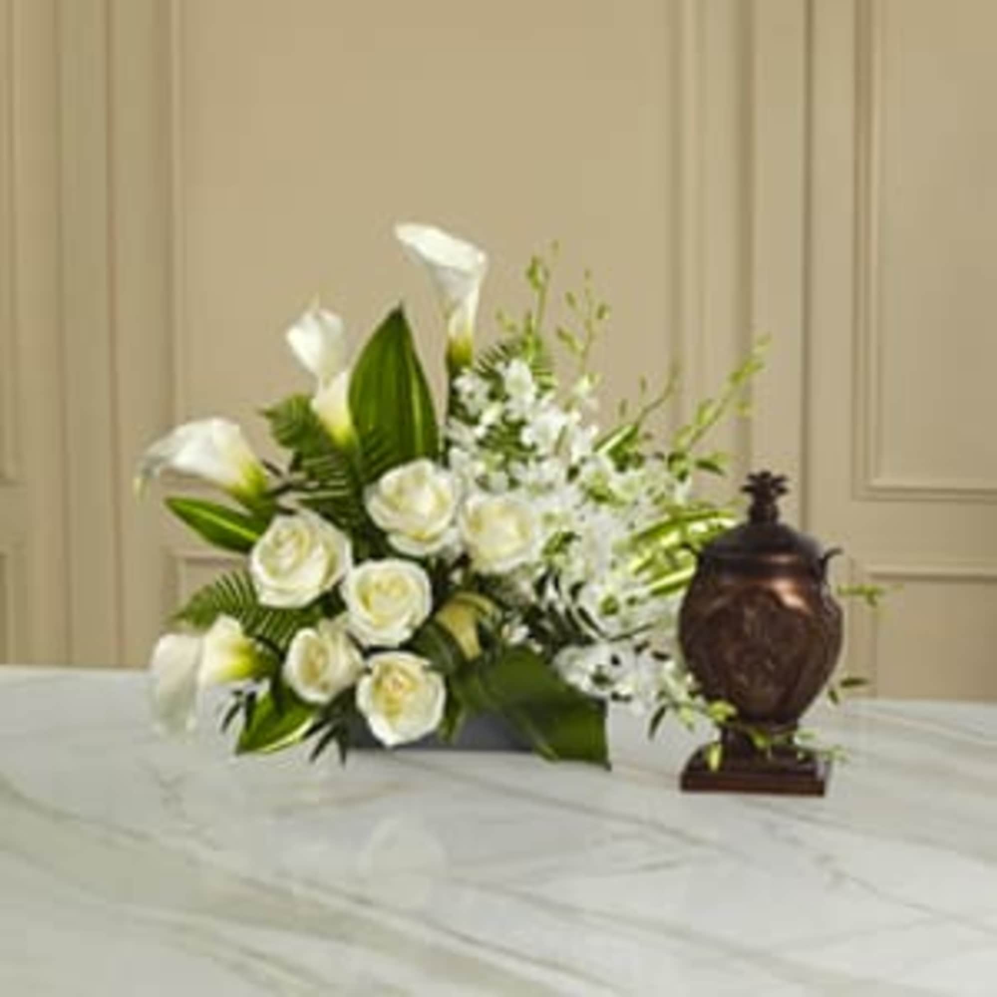 White roses and calla lilies arranged with greenery beside a bronze urn