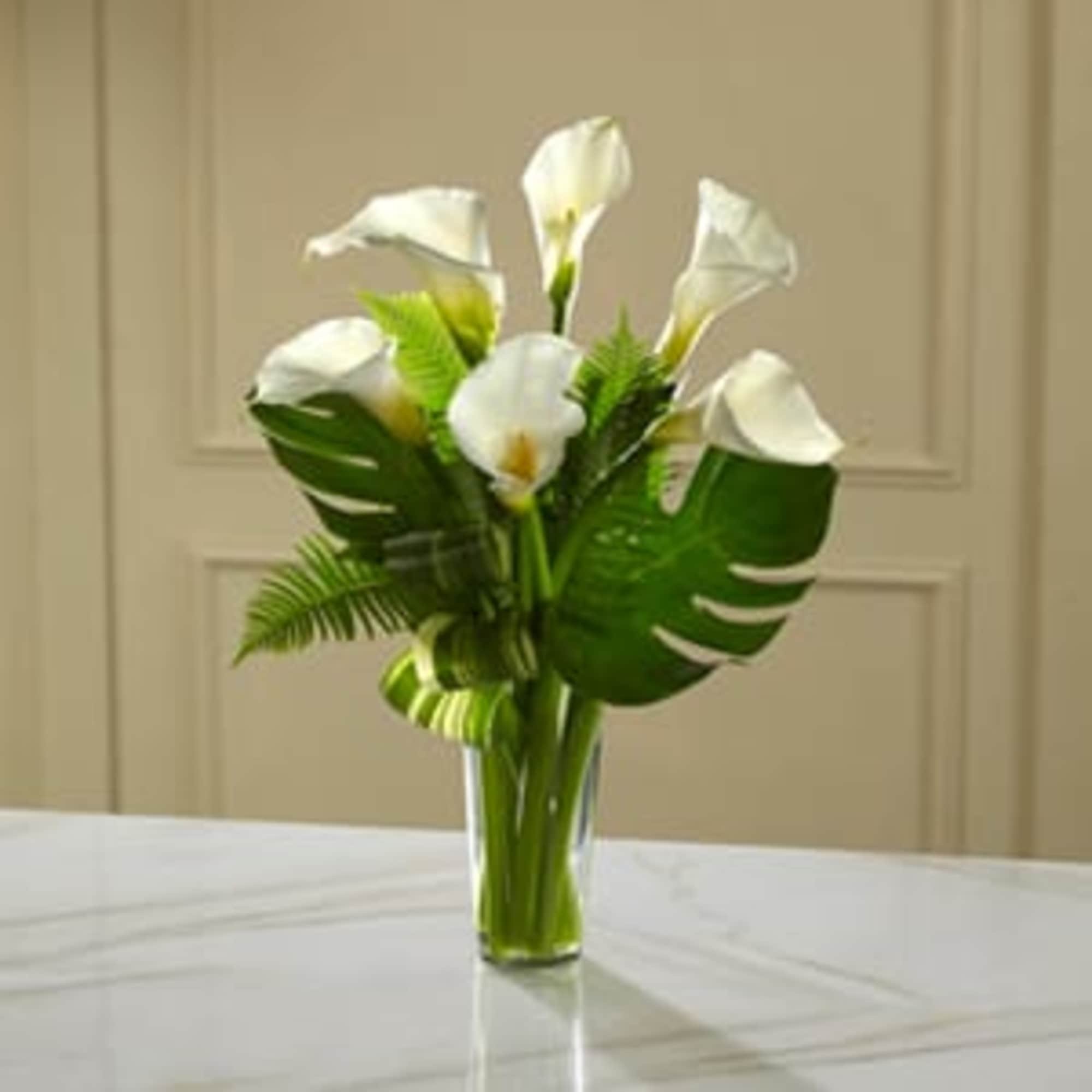 White calla lilies in a clear glass vase with tropical leaves