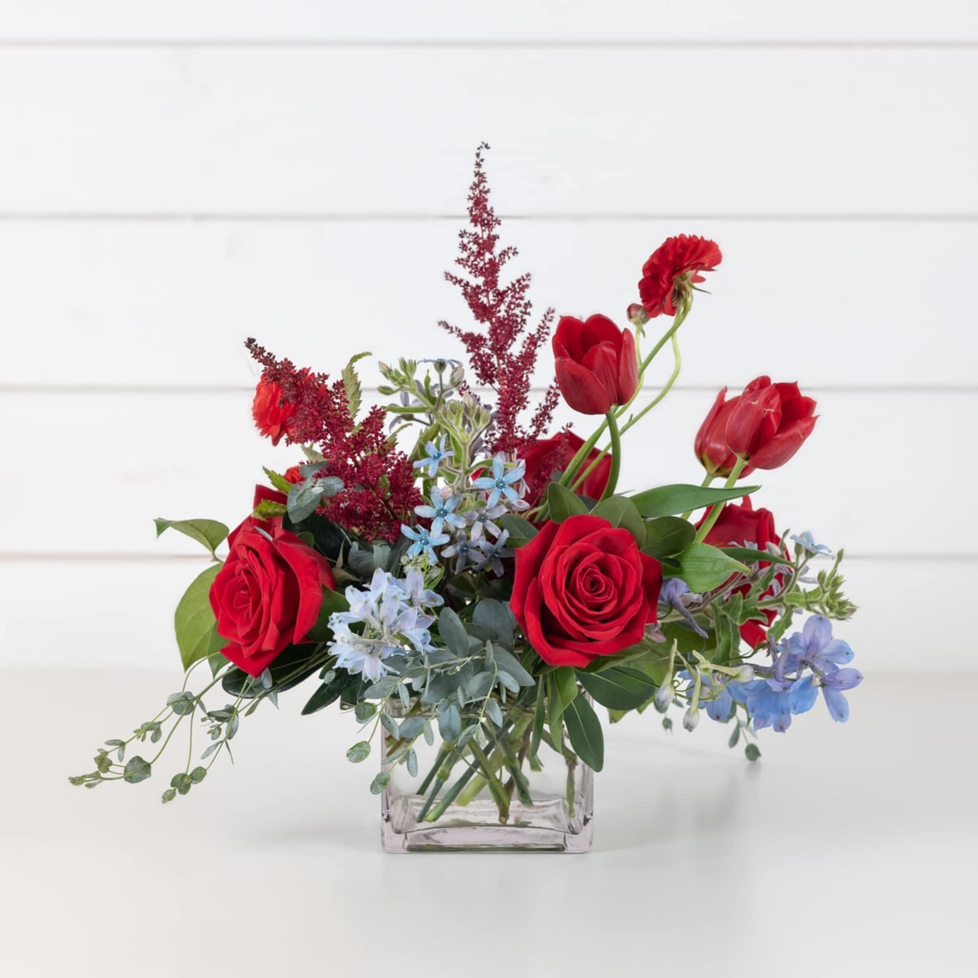Red roses and tulips in a clear square vase with blue flowers