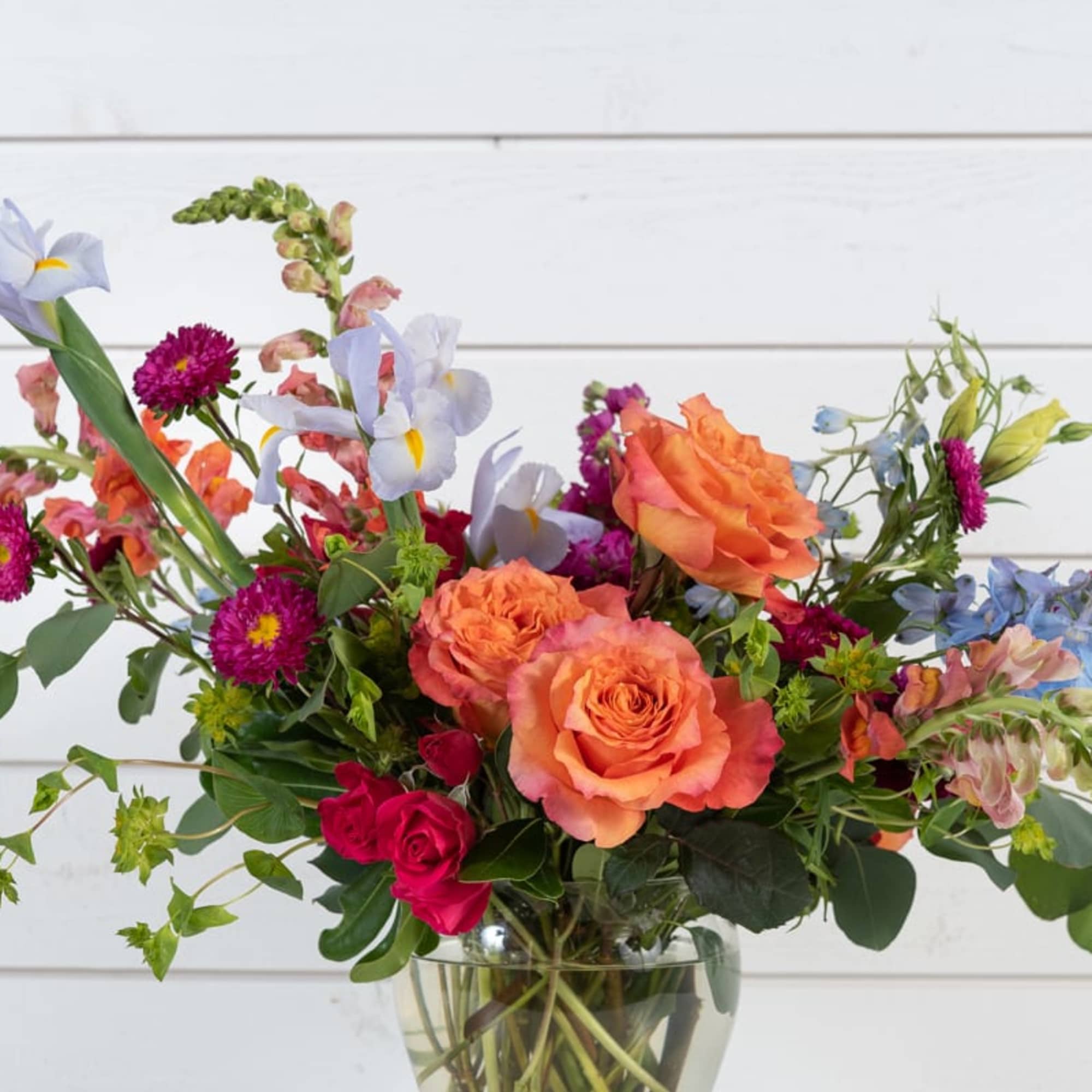 Mixed bouquet of orange and pink roses with blue and purple flowers in a glass vase