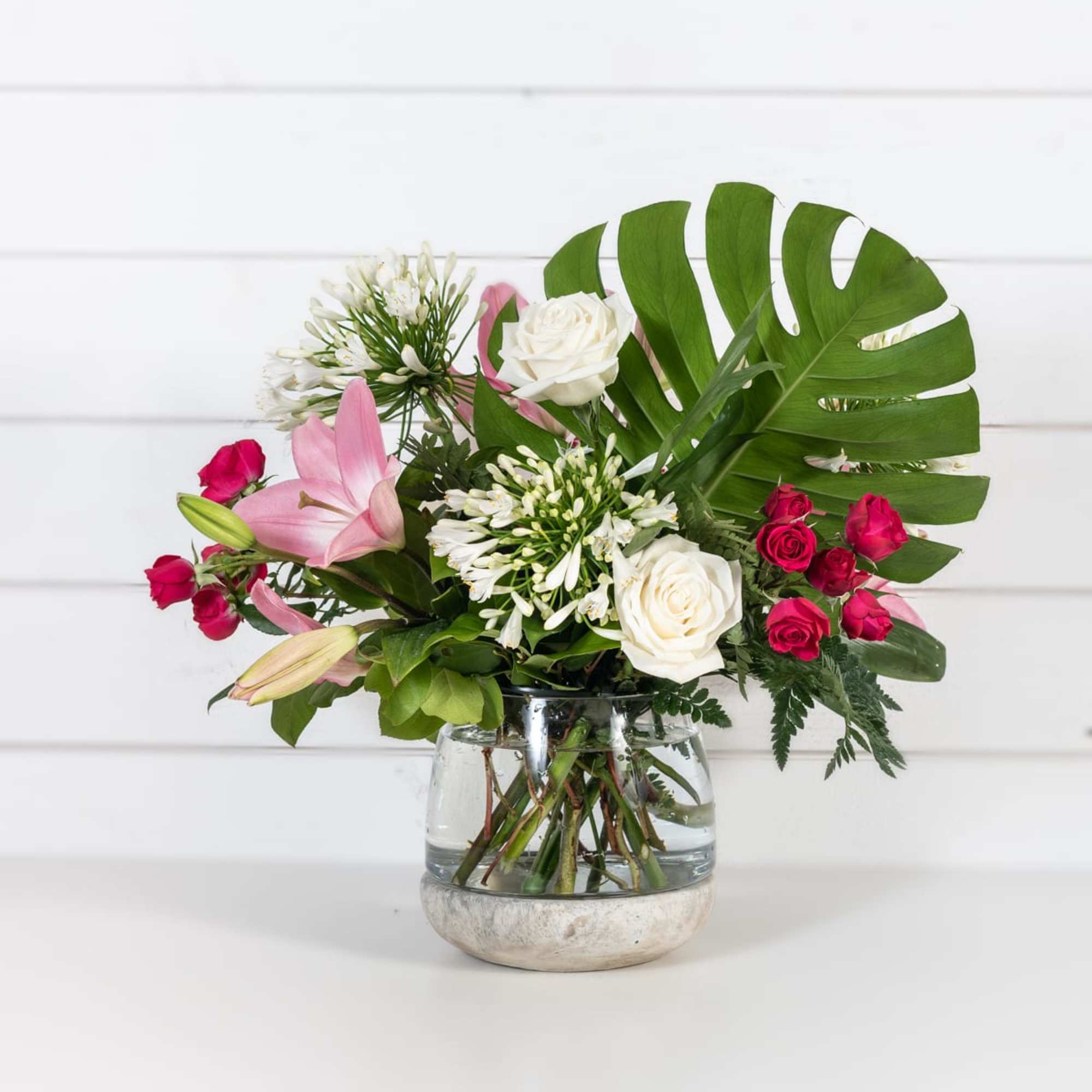 Pink lilies and white roses in a glass vase with tropical leaves