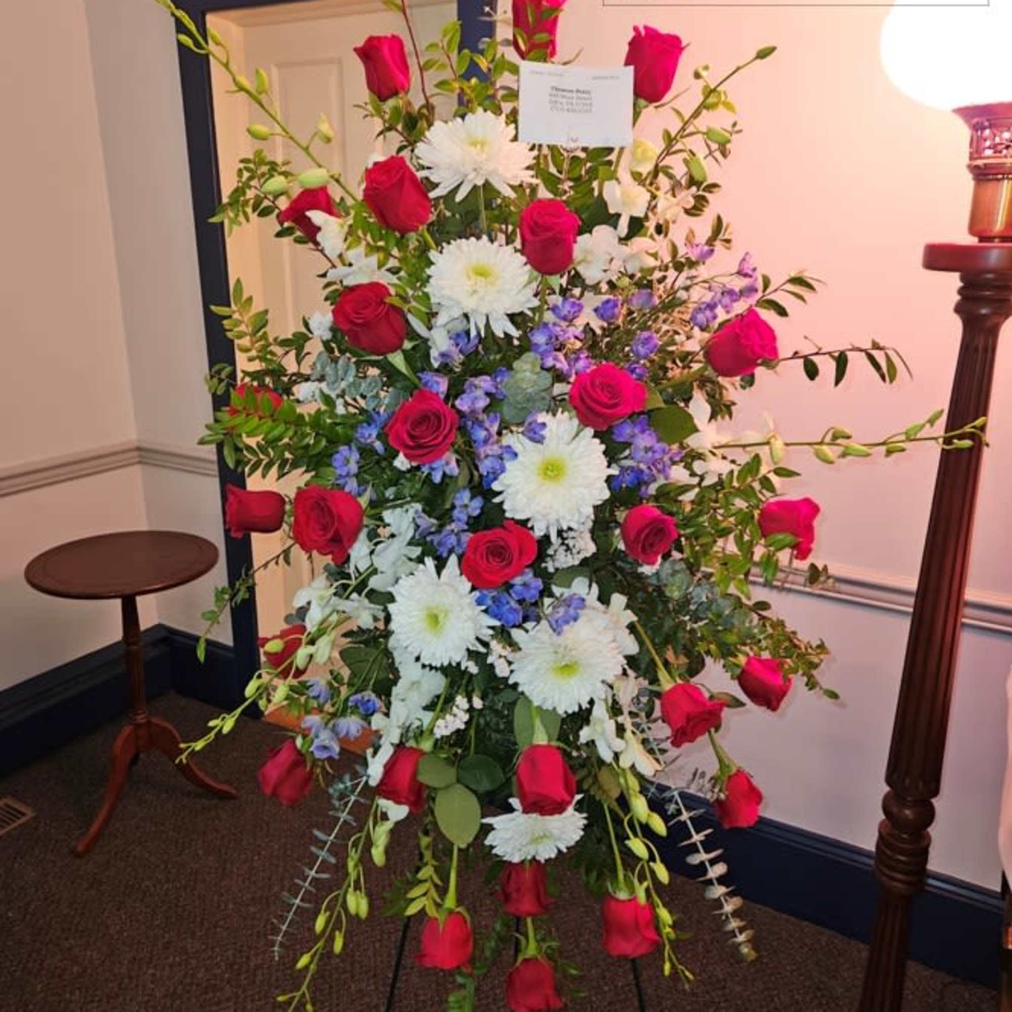 Standing spray of red roses, white mums, and purple flowers on an easel