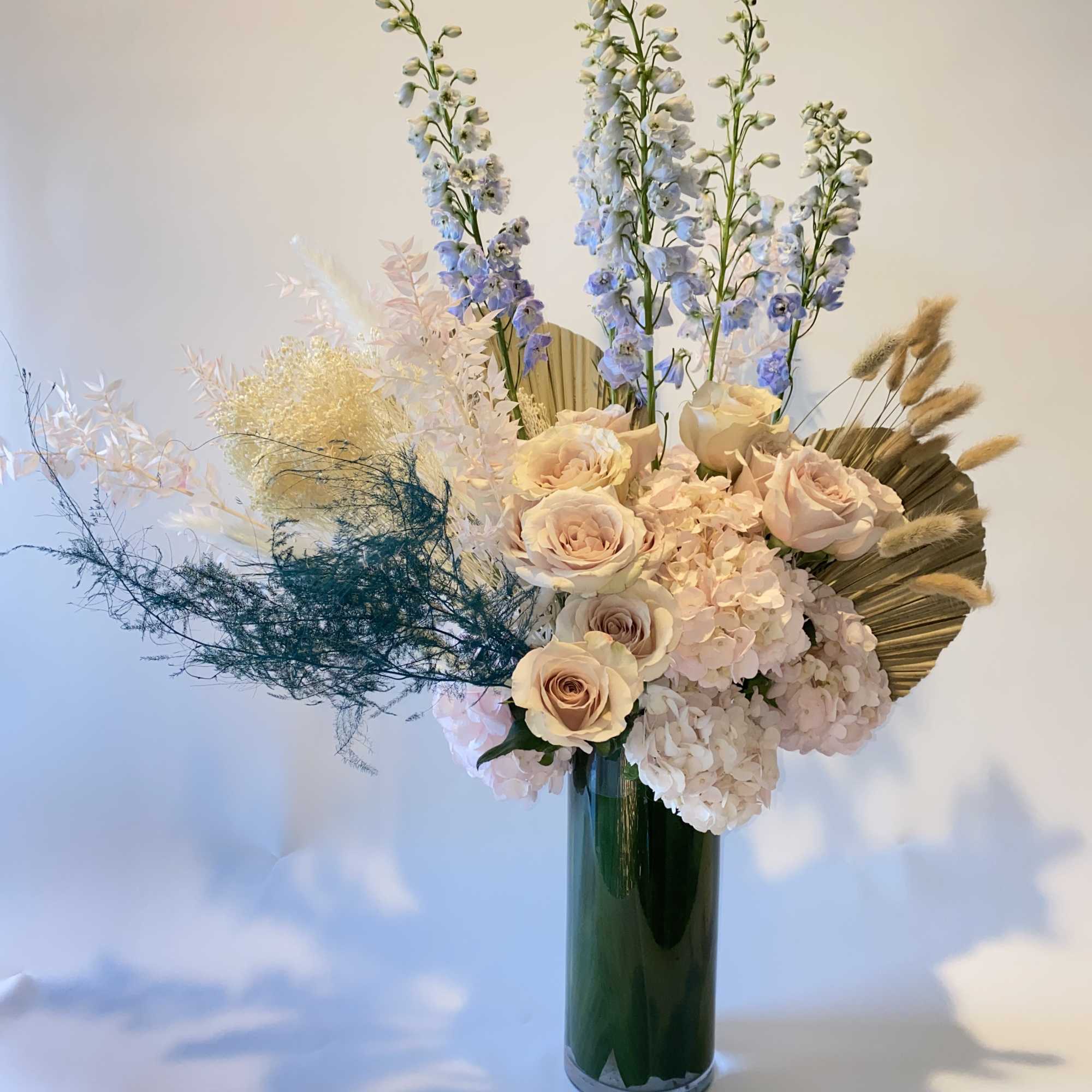 Tall floral arrangement with pale roses, hydrangeas, and blue delphinium in a green vase