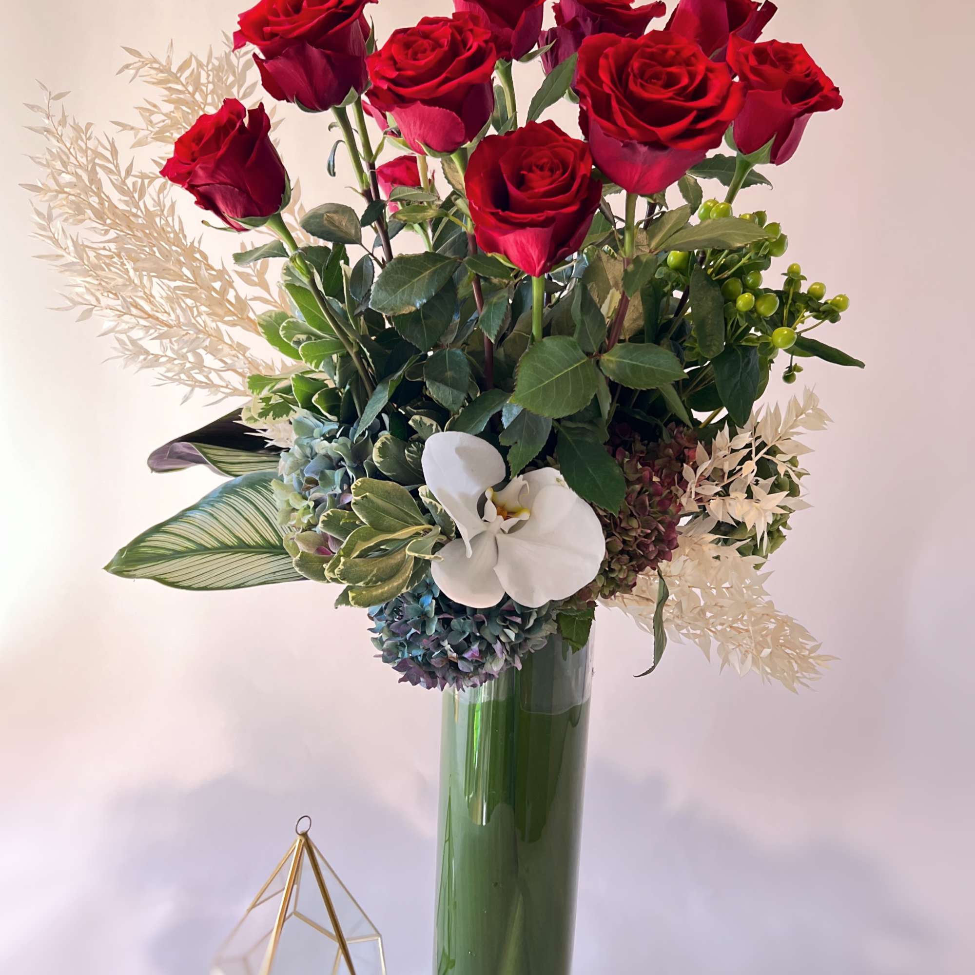 Red roses arranged in a tall green glass vase with white orchid and hydrangea accents