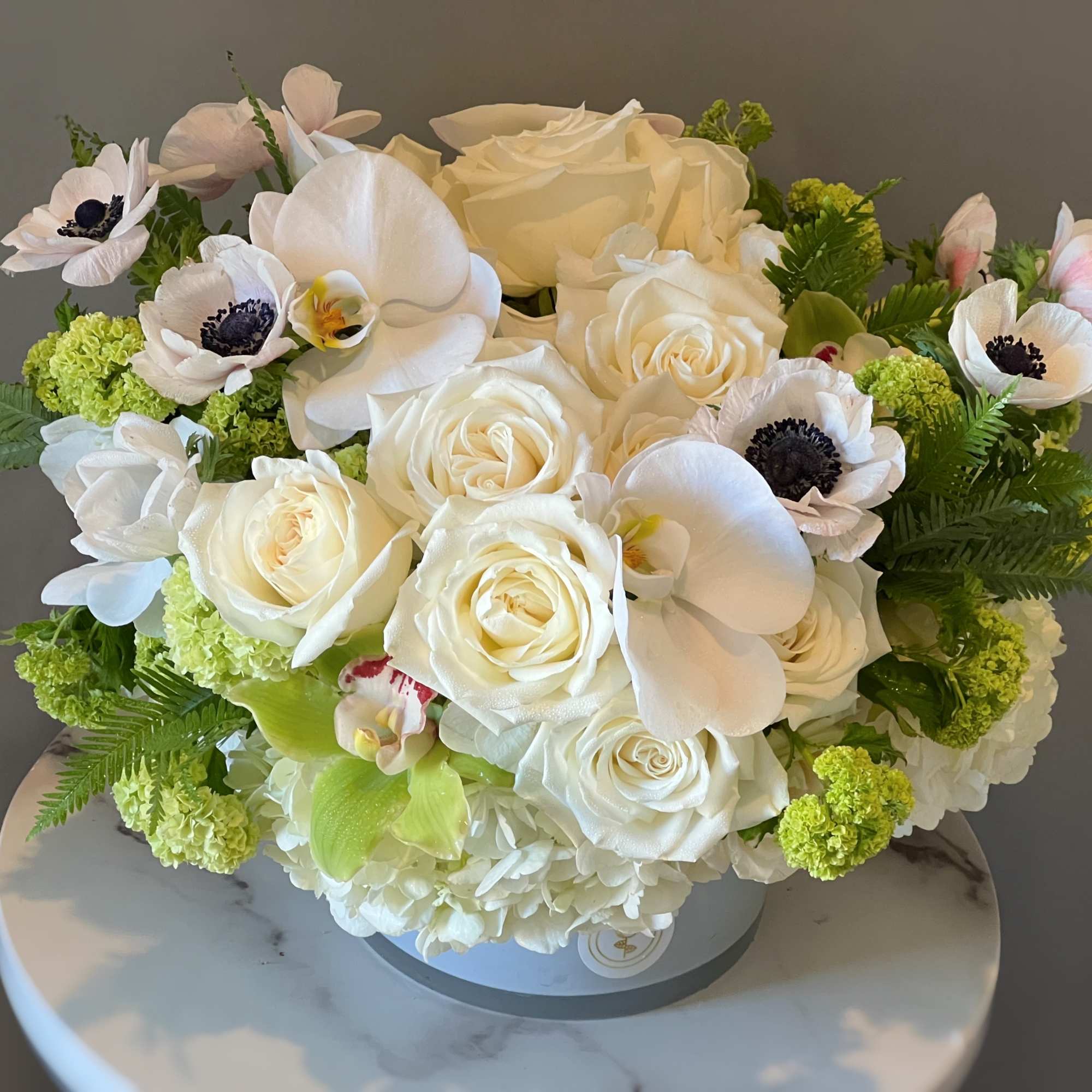 White roses and orchids arranged in a round box with green accents