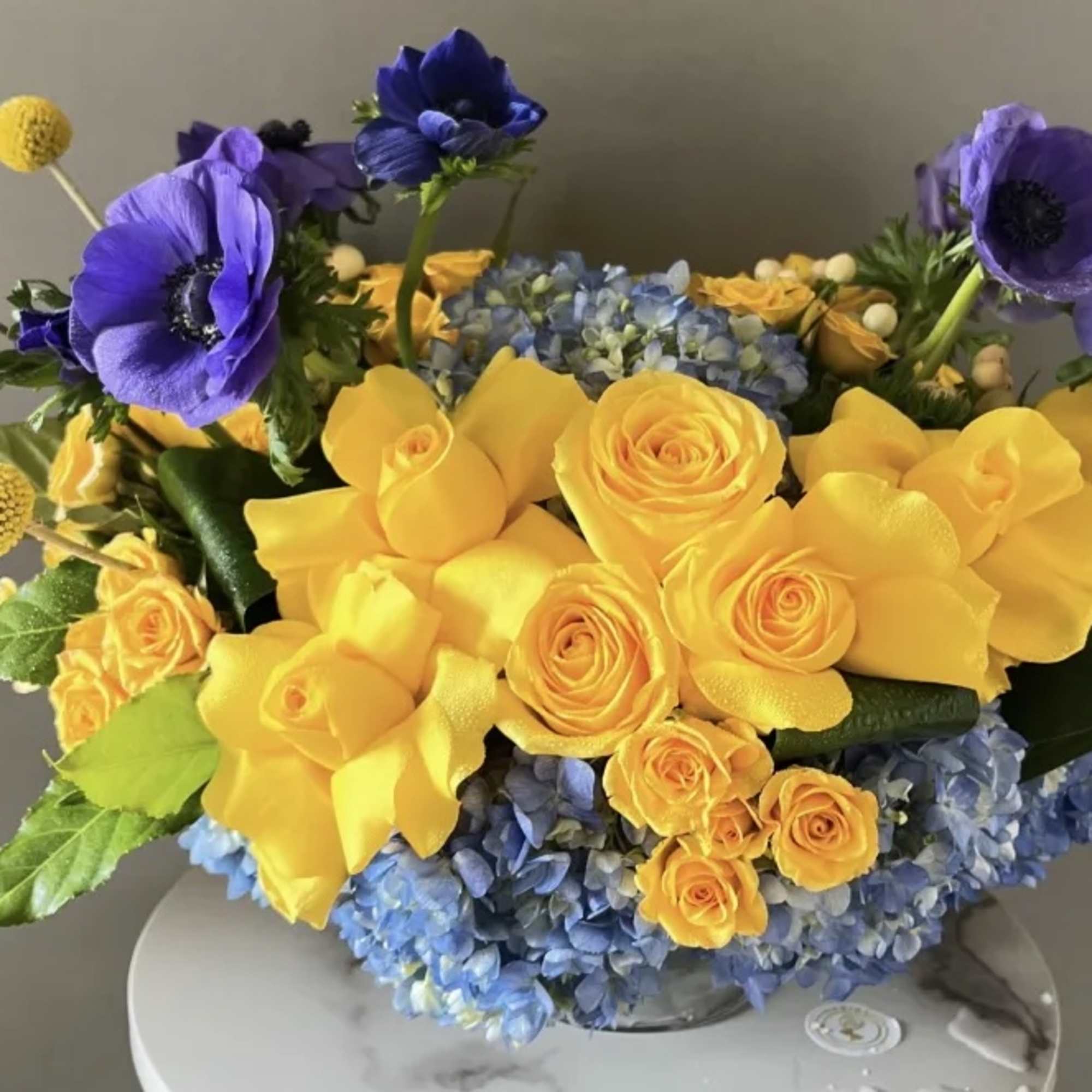 Bouquet of yellow roses, blue hydrangeas, and purple anemones in a glass vase