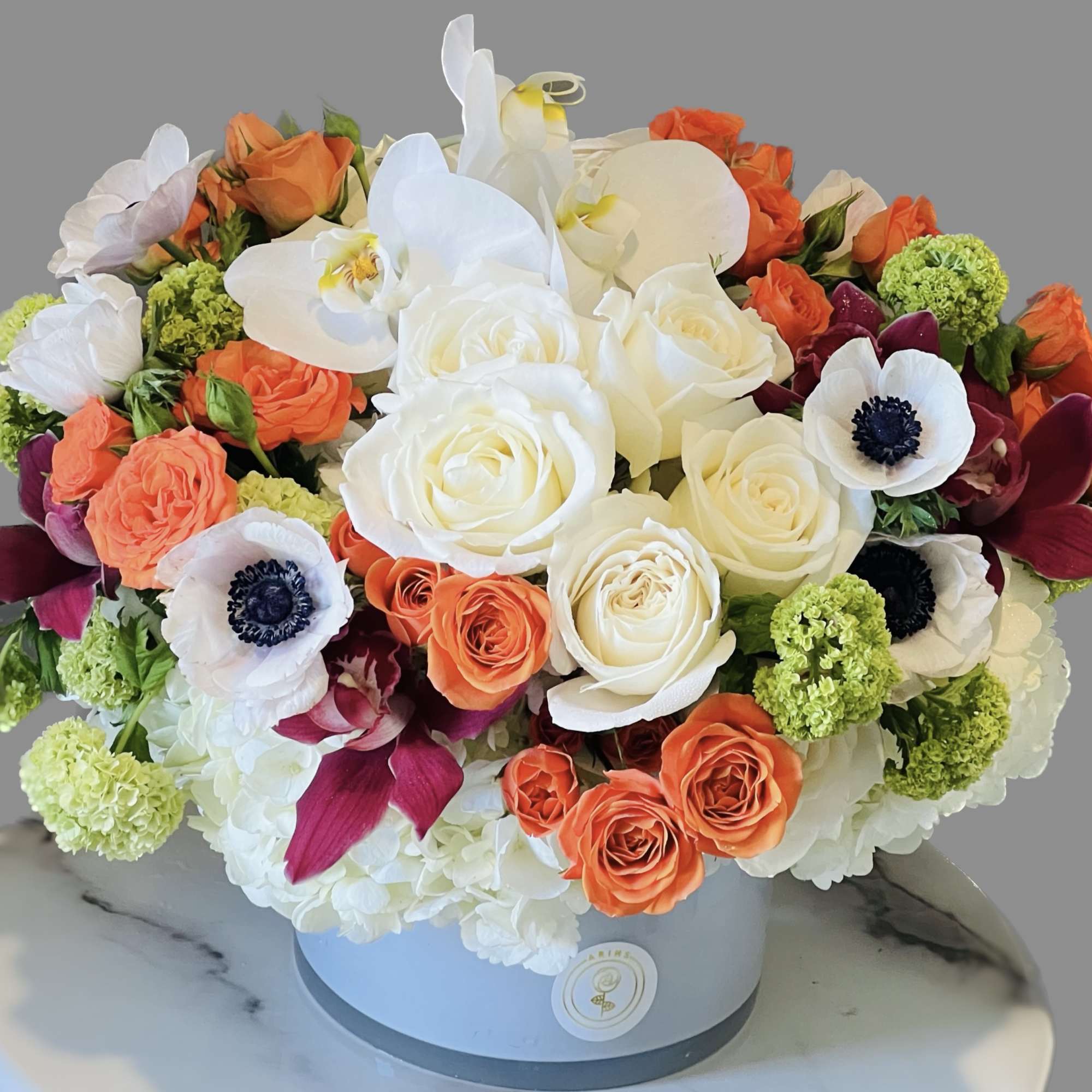 Round bouquet of white orchids, roses, and orange blooms in a hatbox.