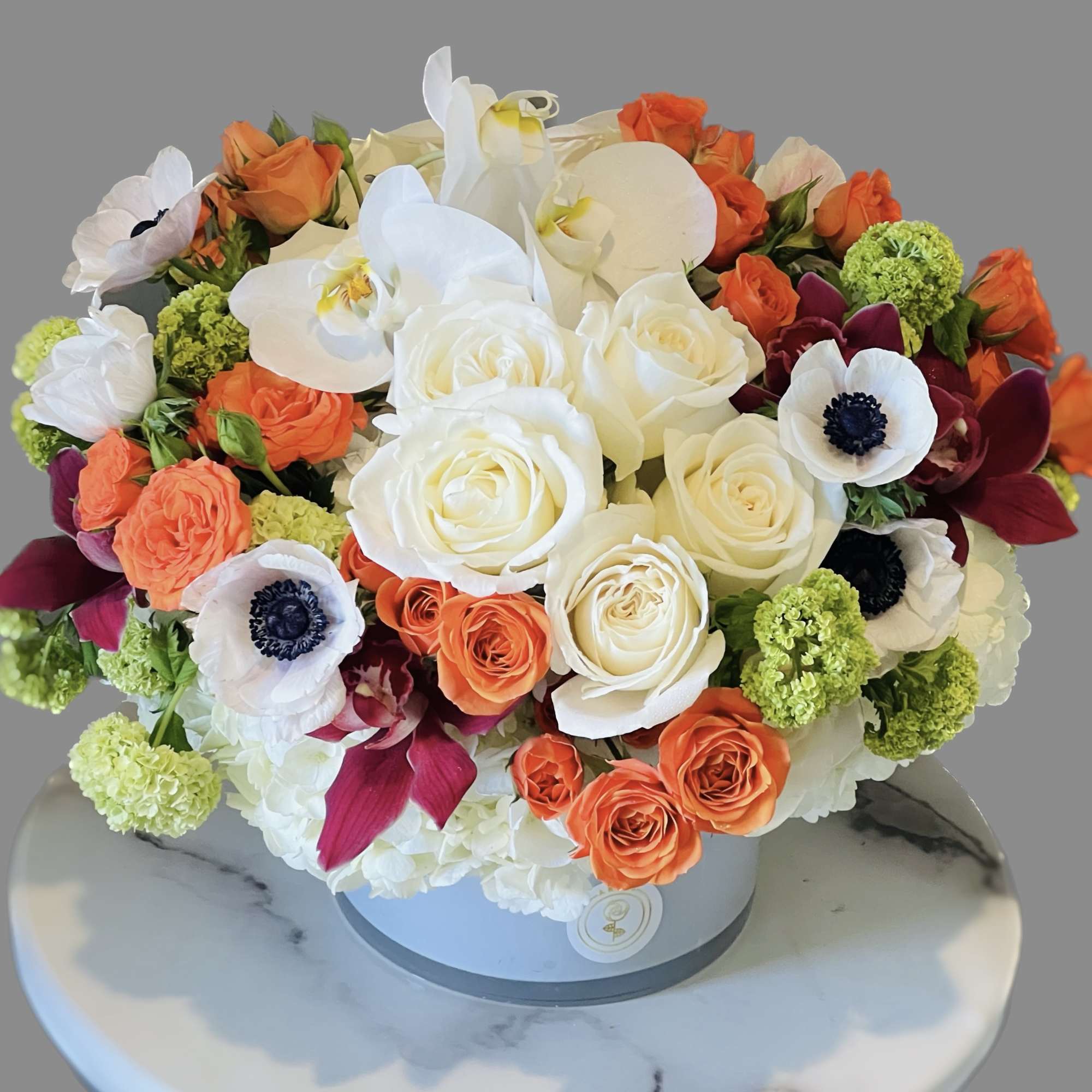 Round bouquet of white orchids, roses, and orange blooms in a gray box