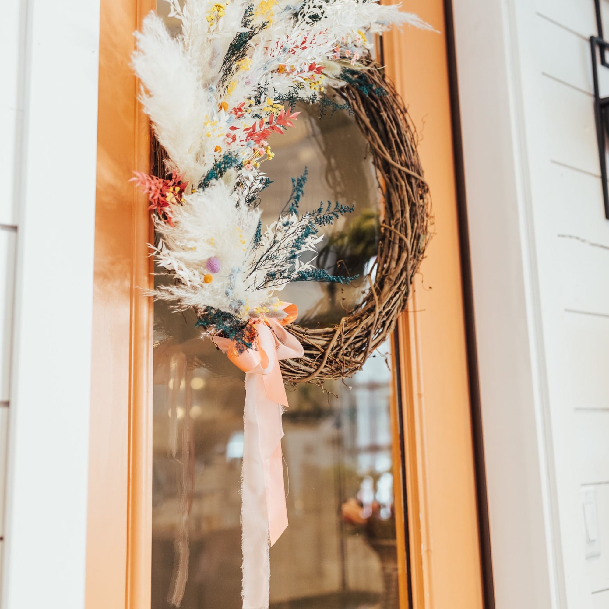 Asymmetrical dried floral wreath with white plumes and pastel accents on a peach front door.
