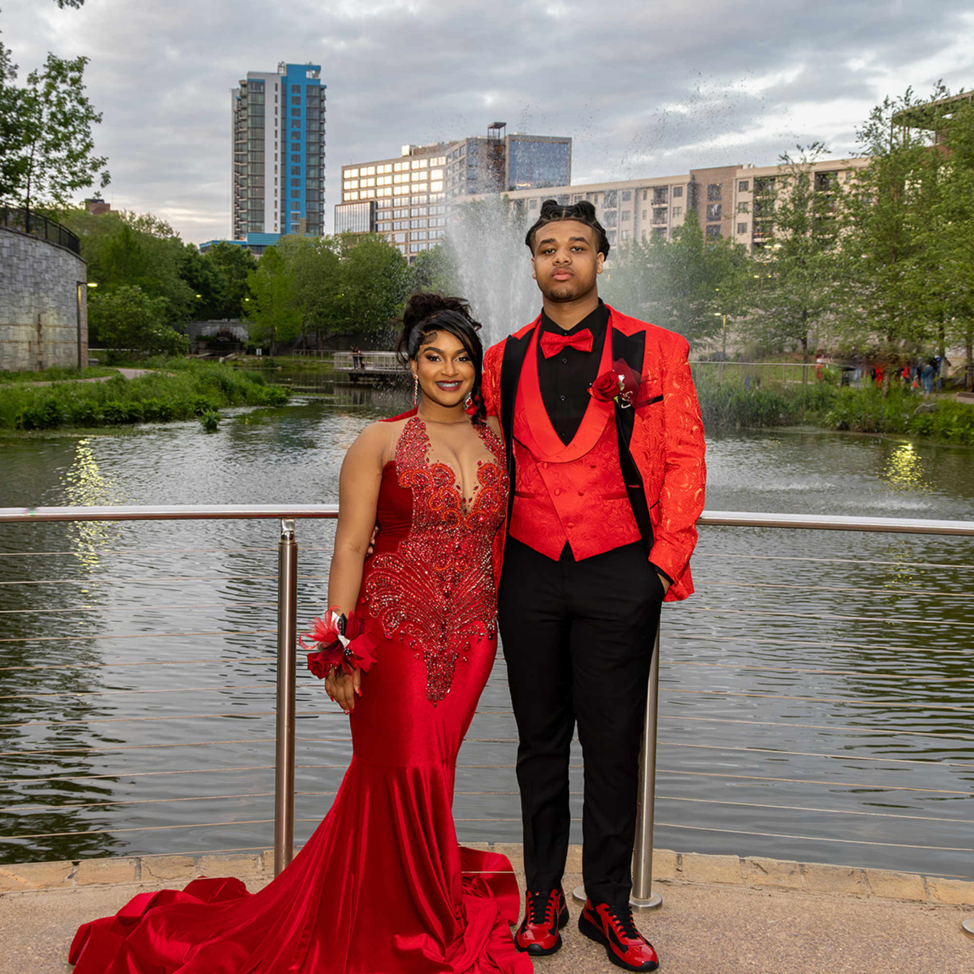A young couple dressed in red formalwear stands by a waterfront fountain.