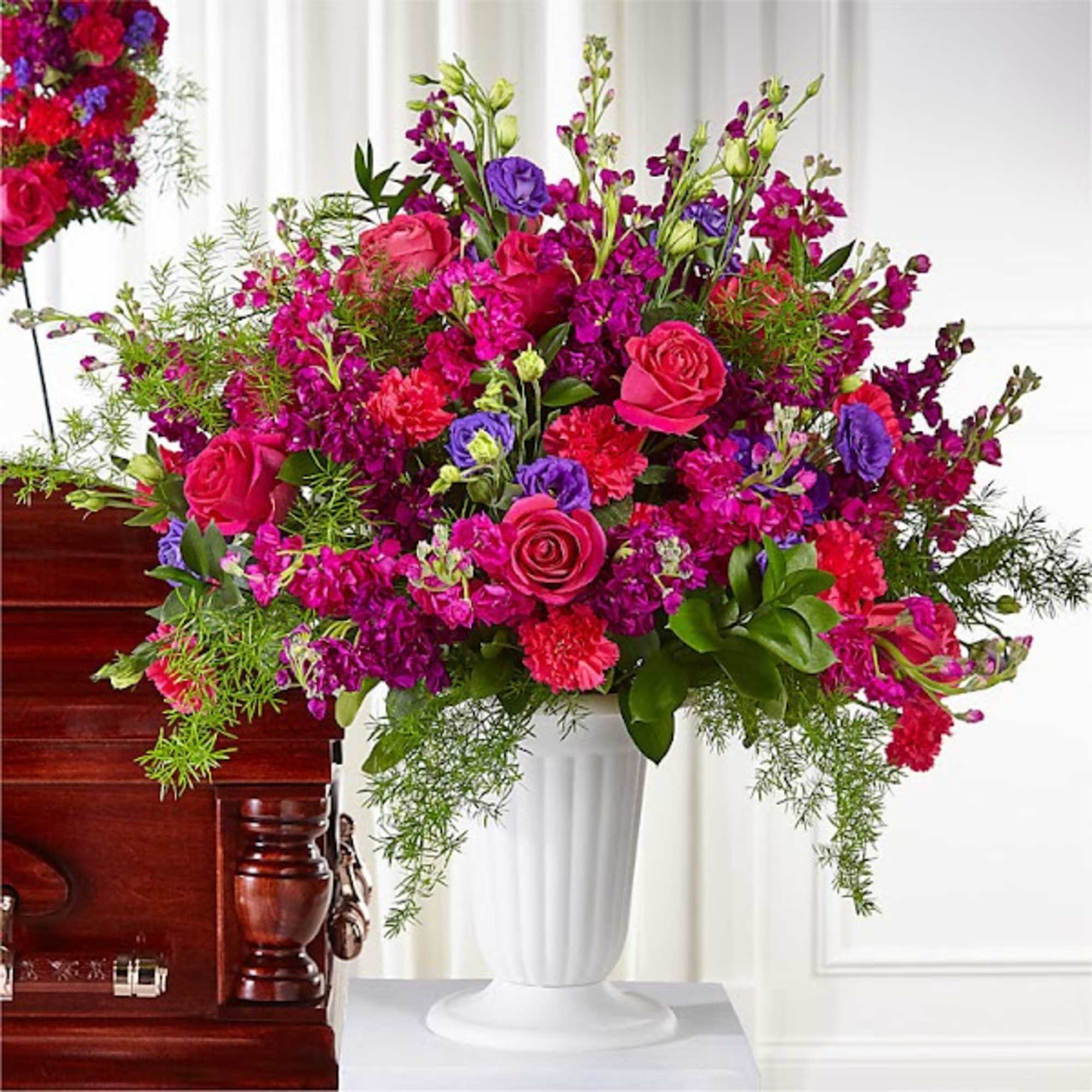 Large pink and purple floral arrangement in a white vase beside a casket