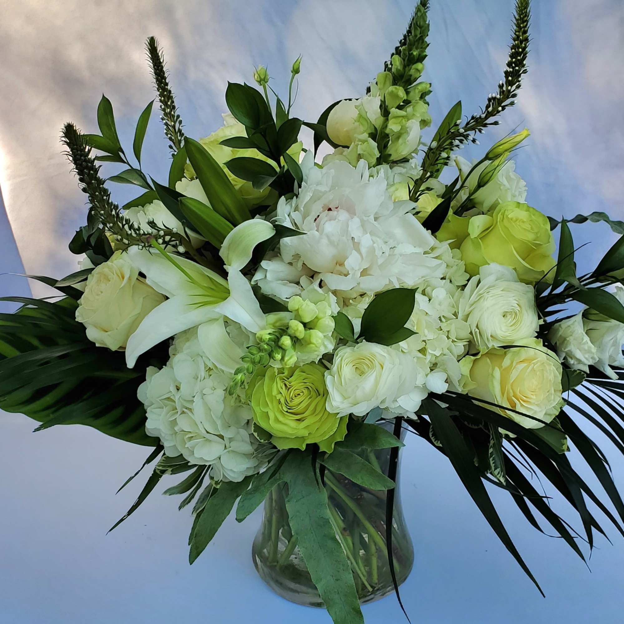 Tall white and cream floral arrangement with lilies, roses, and hydrangeas in a clear glass vase