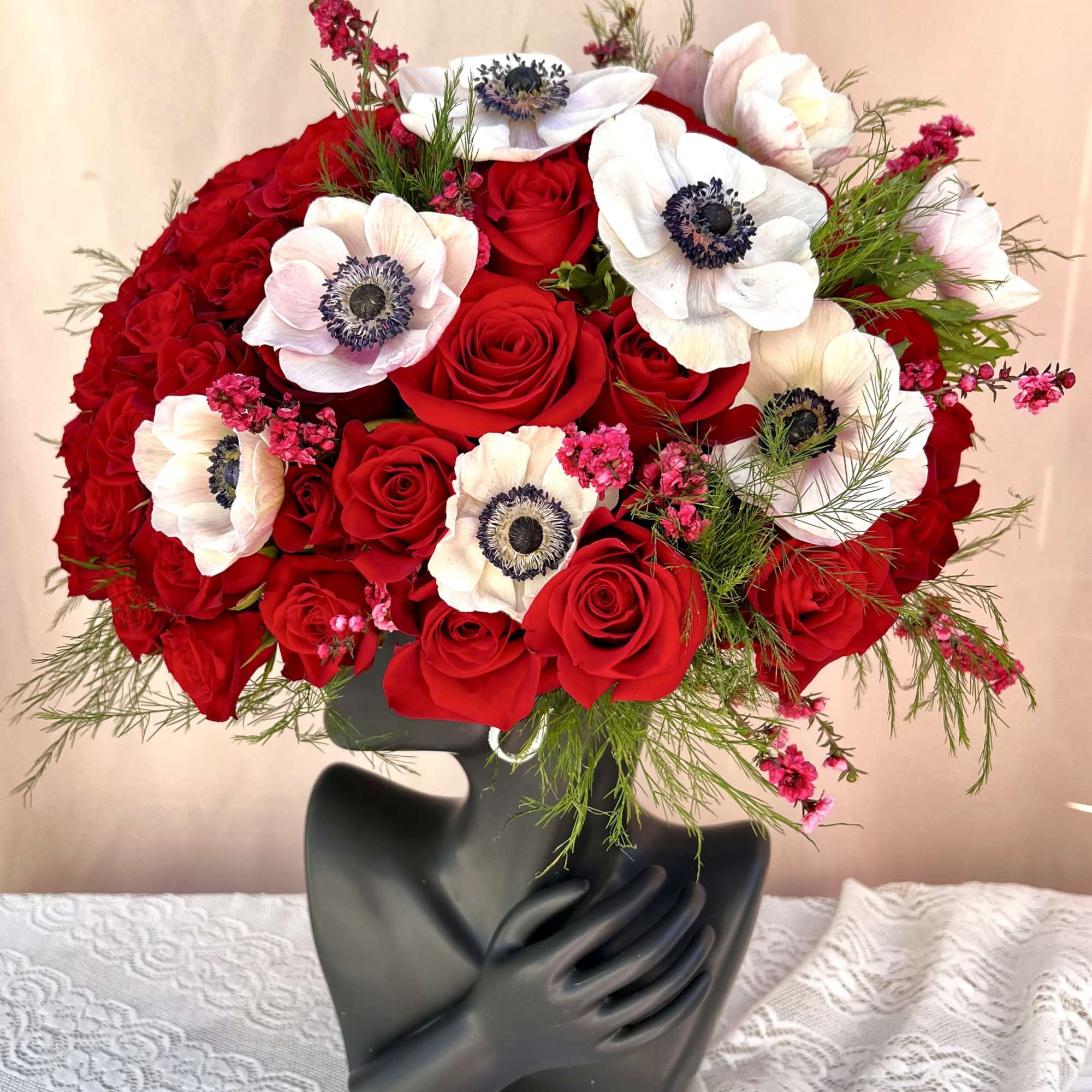Red rose and white anemone arrangement in a black sculptural face vase on a lace-covered table.