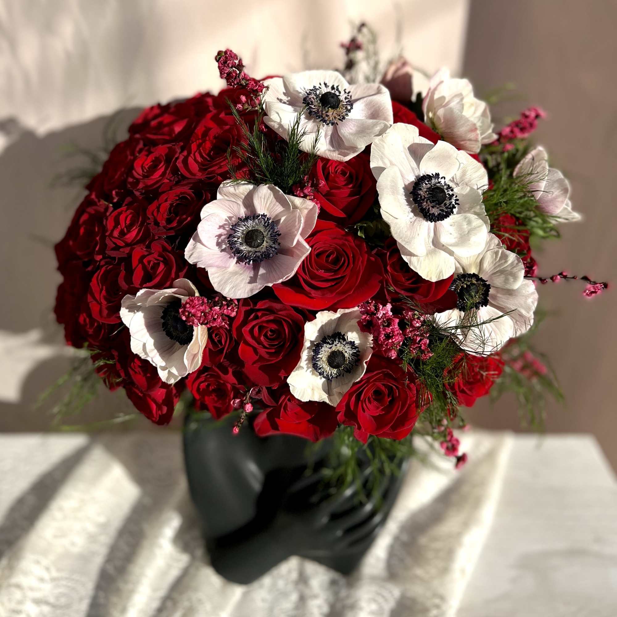 Dome of red roses with white anemones in a matte black sculptural vase on a light surface.